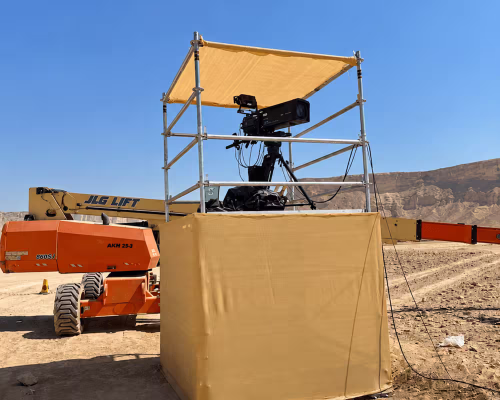 Scaffolding camera platform with protective canopy and JLG lift at an outdoor construction site.