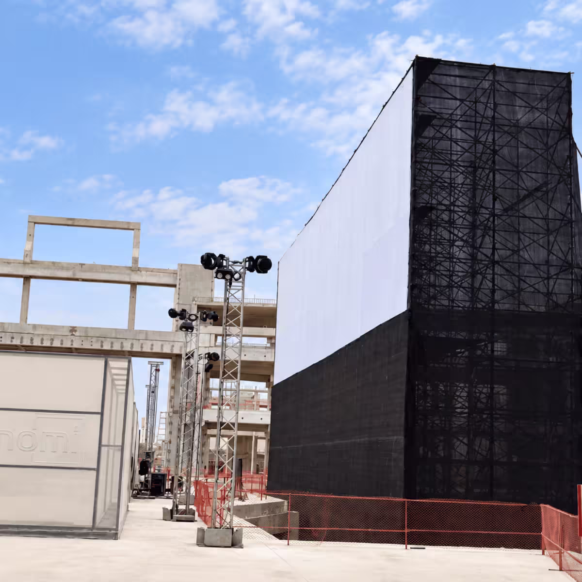 Large temporary event scaffolding structure with black mesh screening, a white backdrop, and lighting towers at an outdoor venue.