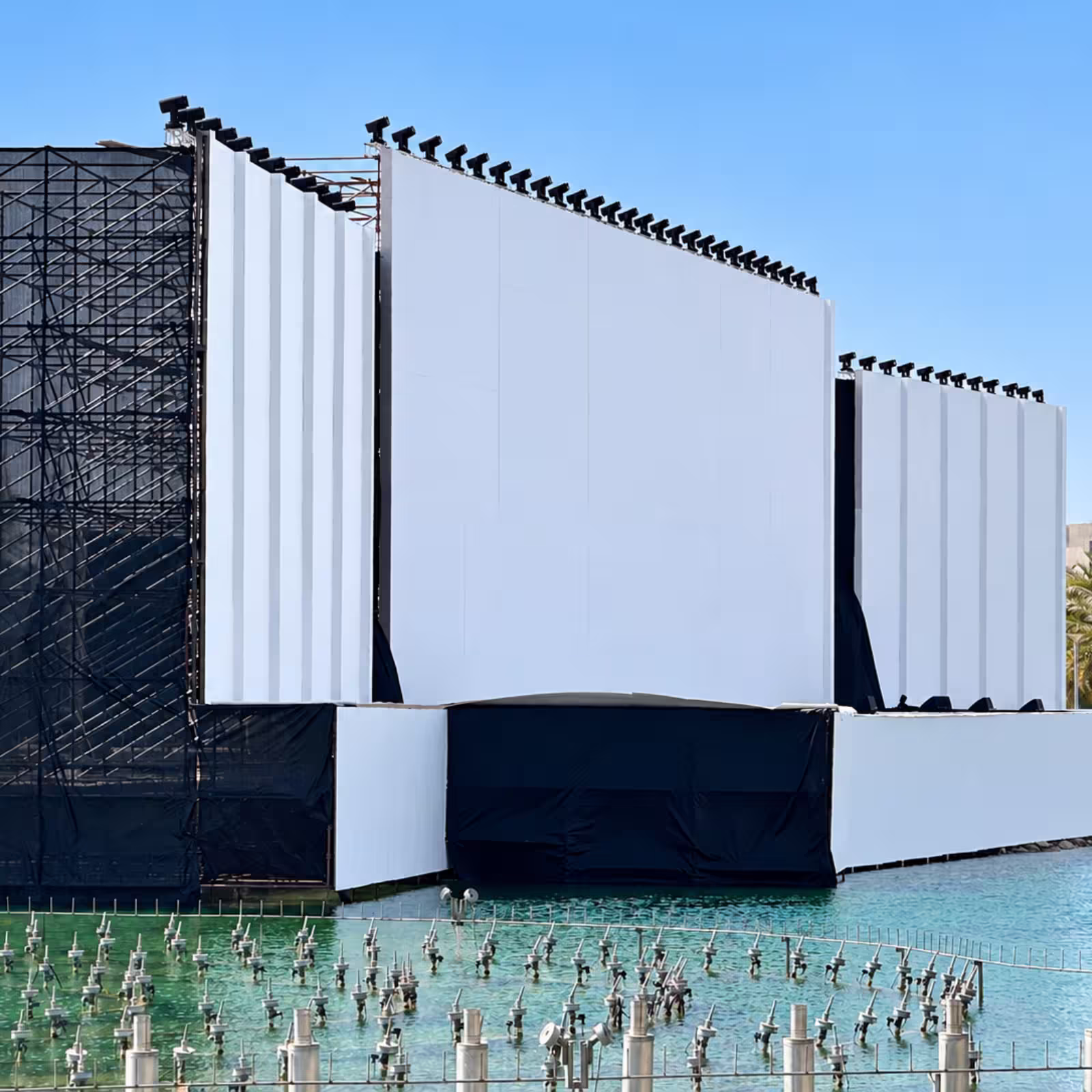 Temporary event structure with large white panels, black scaffolding, and lighting fixtures above a water feature.