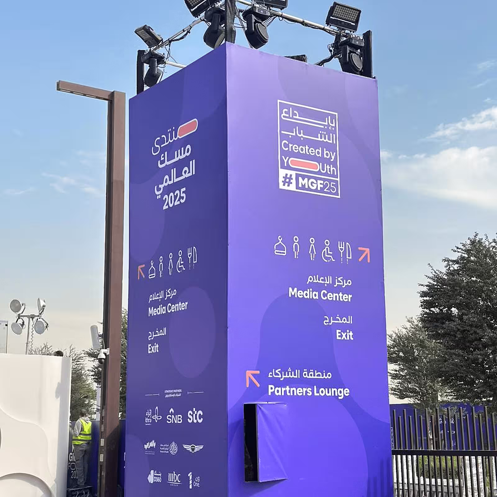 Purple event signage tower with lighting fixtures, directional information, and bilingual Arabic-English branding.