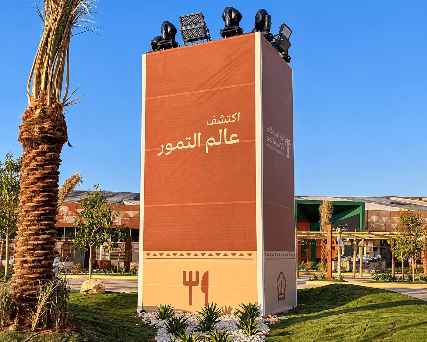 Orange event branding tower with Arabic signage, decorative landscaping, and multiple stage lighting fixtures.