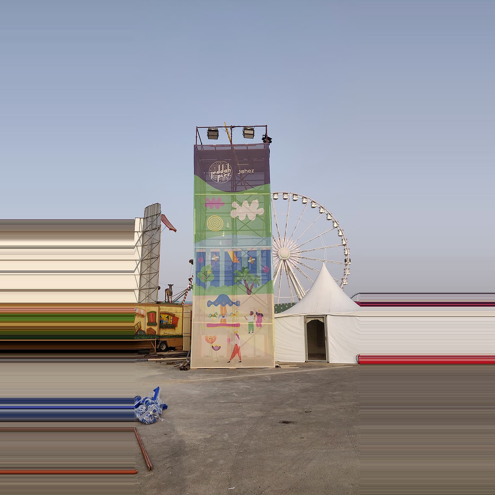 Colorful temporary event tower with printed graphics, stage lighting, a Ferris wheel, and a large event tent in the background.