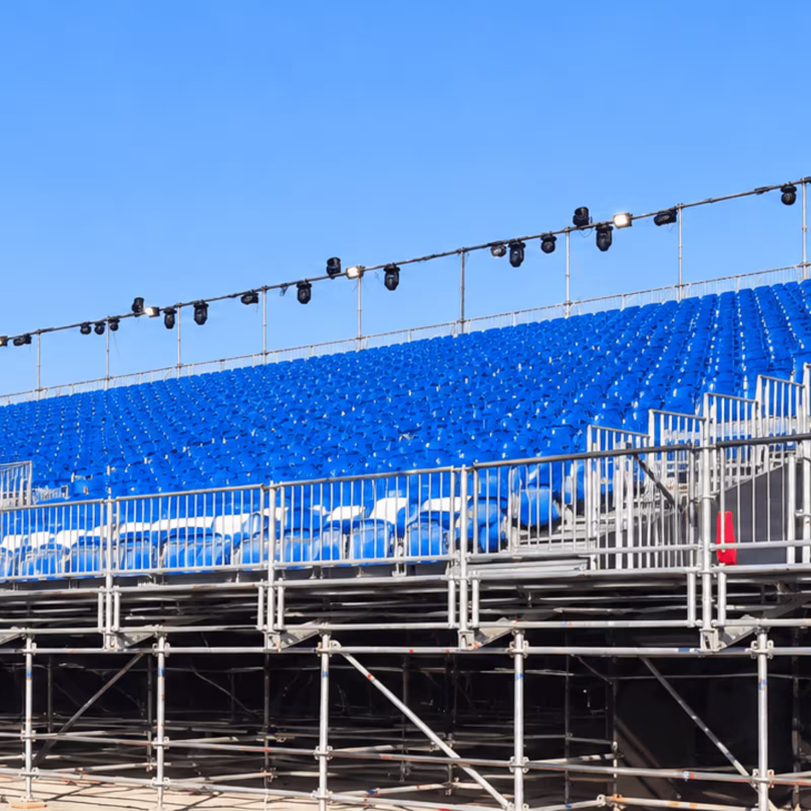Temporary grandstand seating with blue spectator chairs, metal railings, scaffolding support, and event lighting infrastructure.