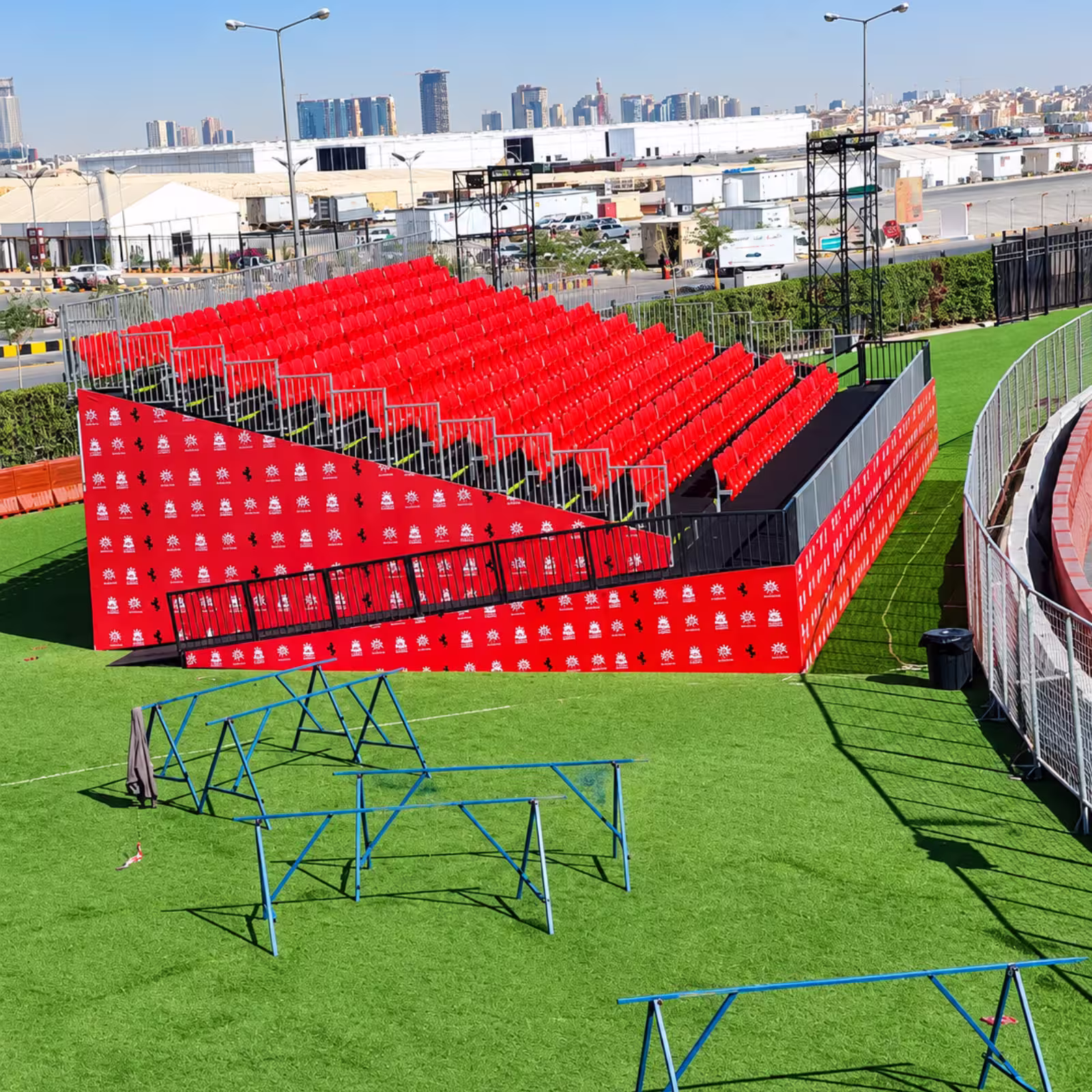 Temporary event grandstand with red spectator seating, safety railings, branded panels, and an artificial turf event area.