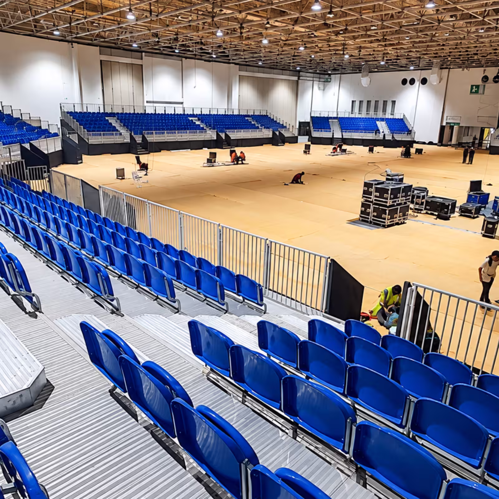 Indoor temporary grandstand seating with blue spectator chairs, aluminum platforms, safety railings, and an open event floor prepared for setup.