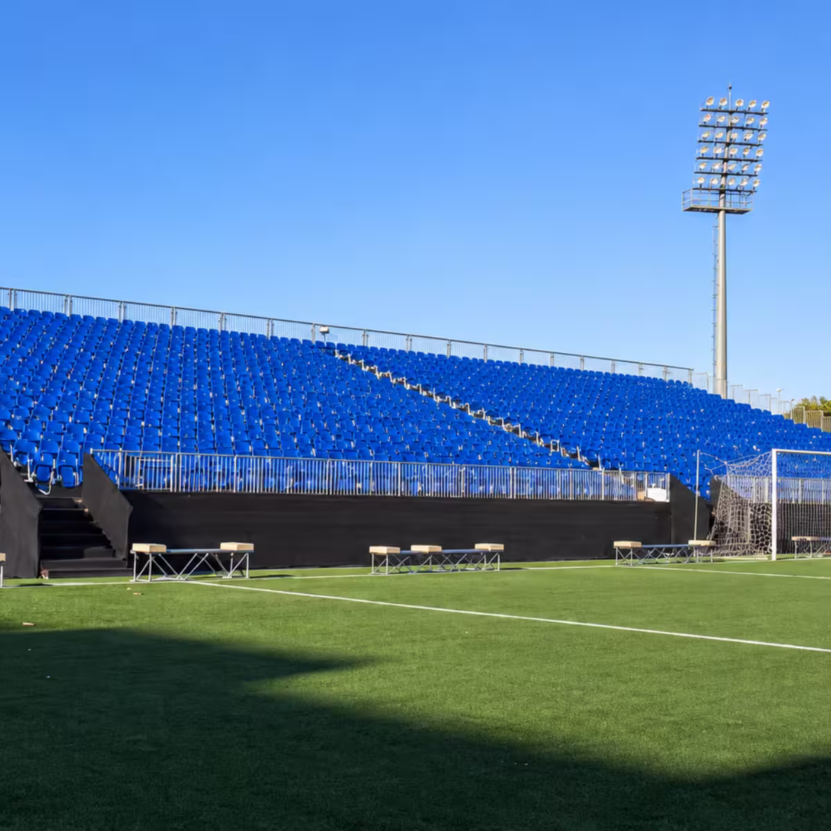 Outdoor temporary grandstand seating with blue spectator chairs, safety railings, artificial turf, and stadium floodlighting.