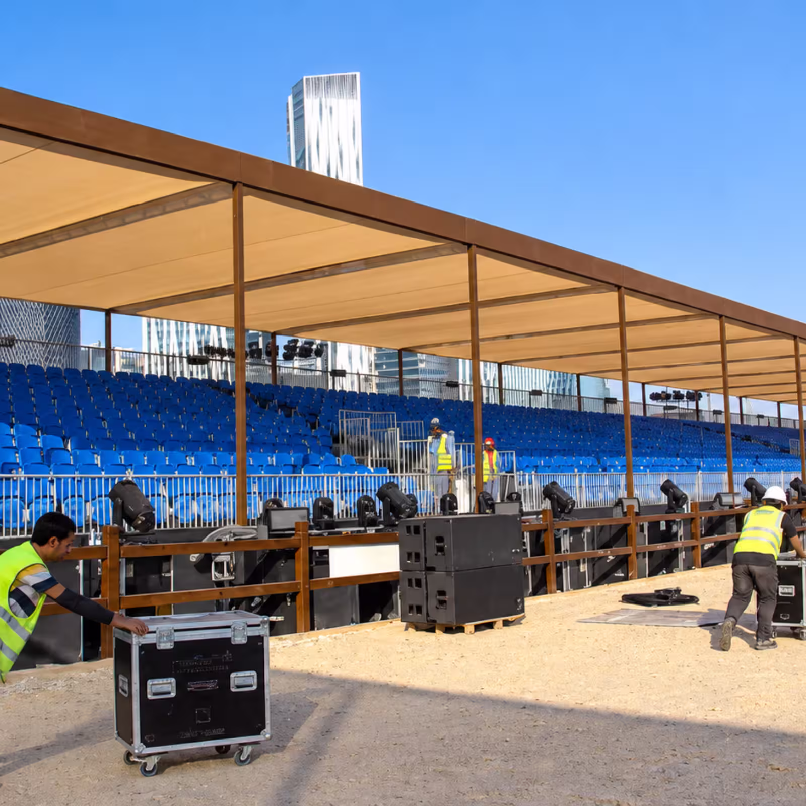 Outdoor temporary grandstand with blue spectator seating, a covered canopy, safety railings, event lighting, and technical equipment being installed.