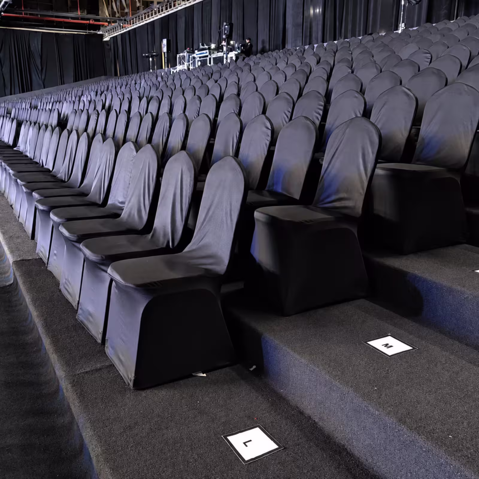 Indoor temporary grandstand seating with rows of black and grey chair covers, tiered platforms, and clearly marked seat positions for an organized event setup.