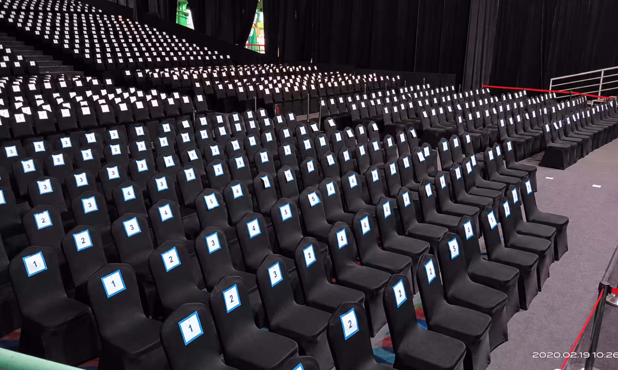 Indoor temporary grandstand seating with black chair covers, numbered reserved seats, tiered rows, and organized aisle access for a large-scale event.
