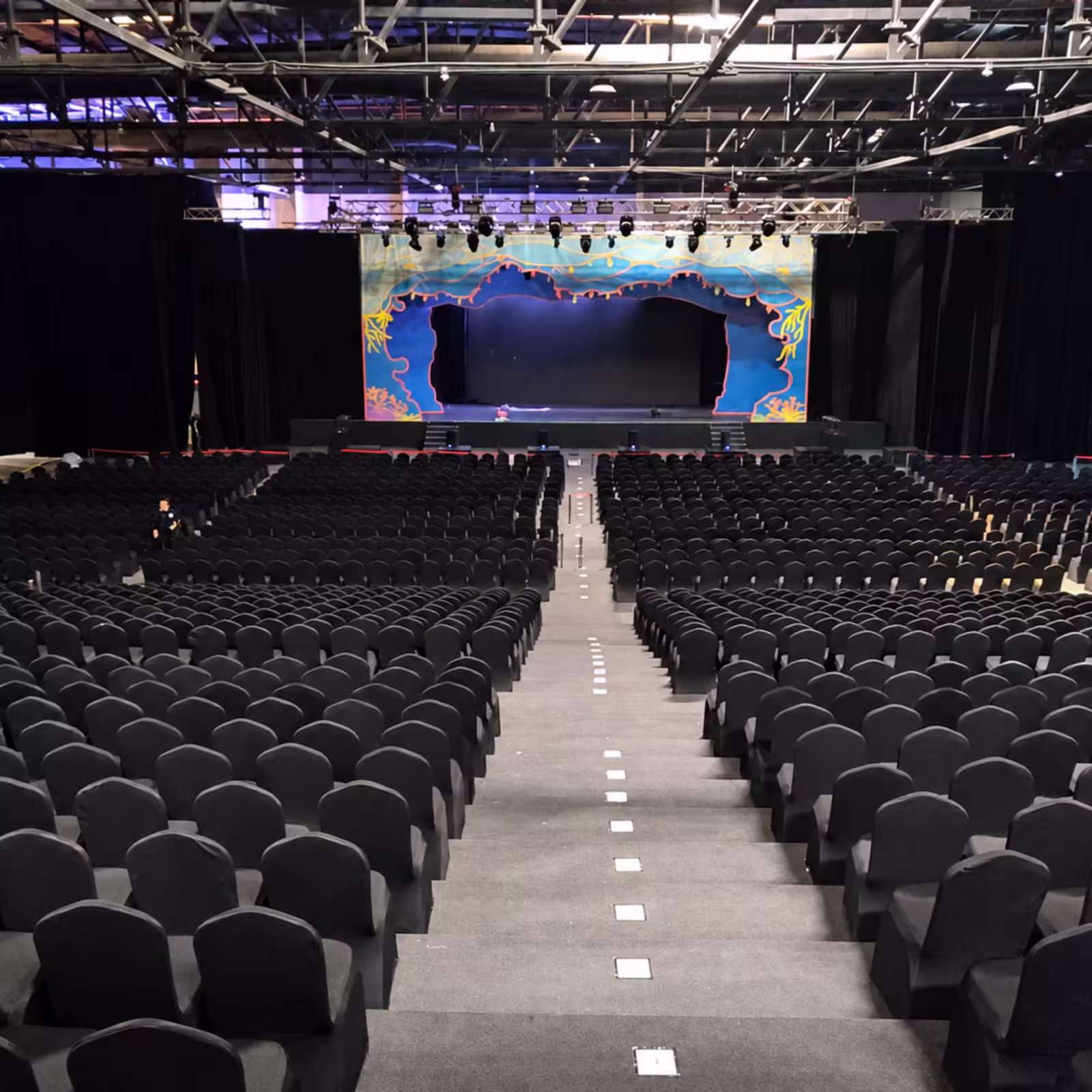 Indoor event auditorium with hundreds of black covered seats arranged in tiered sections, a central aisle, stage lighting, and a large decorated stage backdrop.