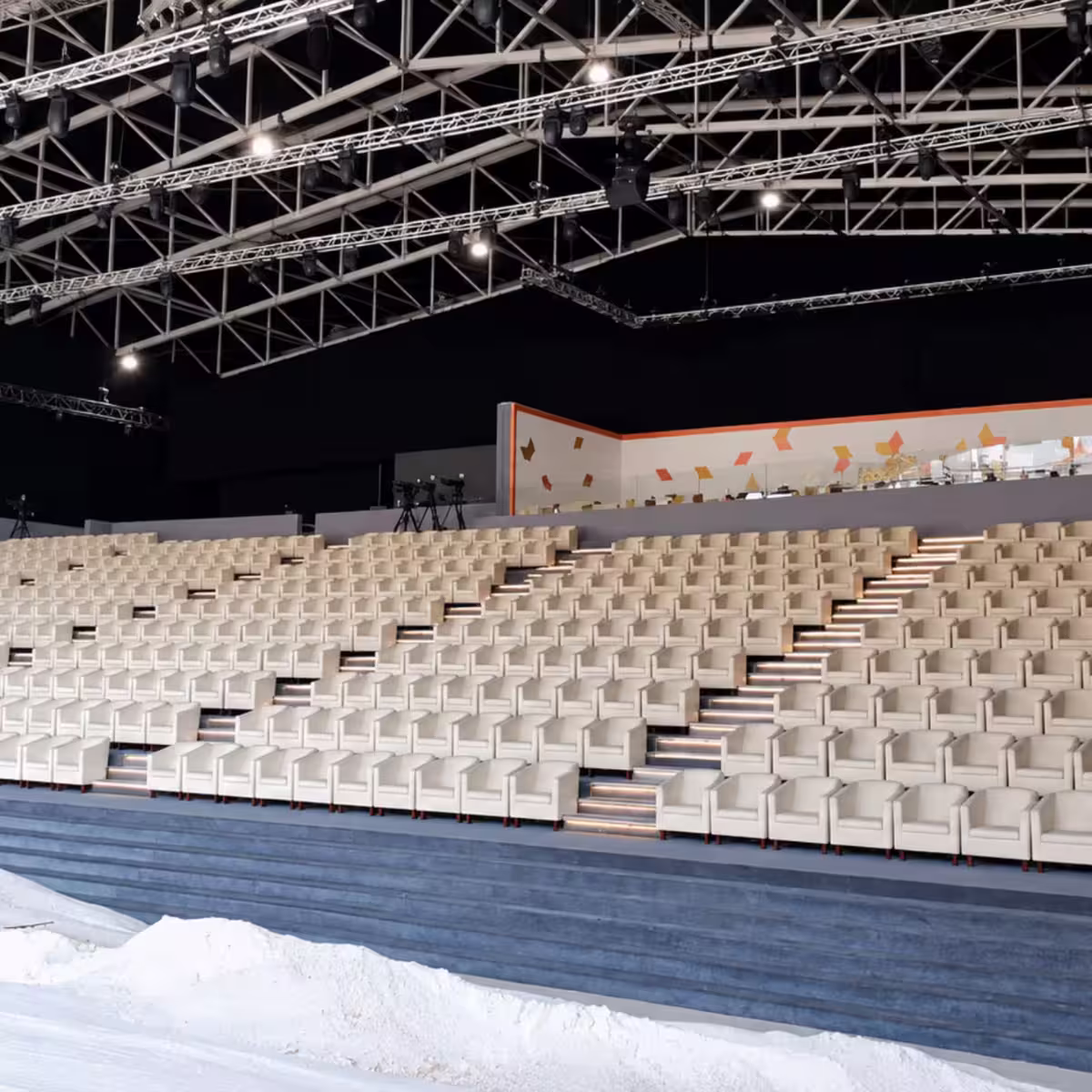 Indoor event grandstand featuring rows of white lounge-style seats arranged in tiers, illuminated stair aisles, and overhead truss lighting for a large-scale event or exhibition setup.