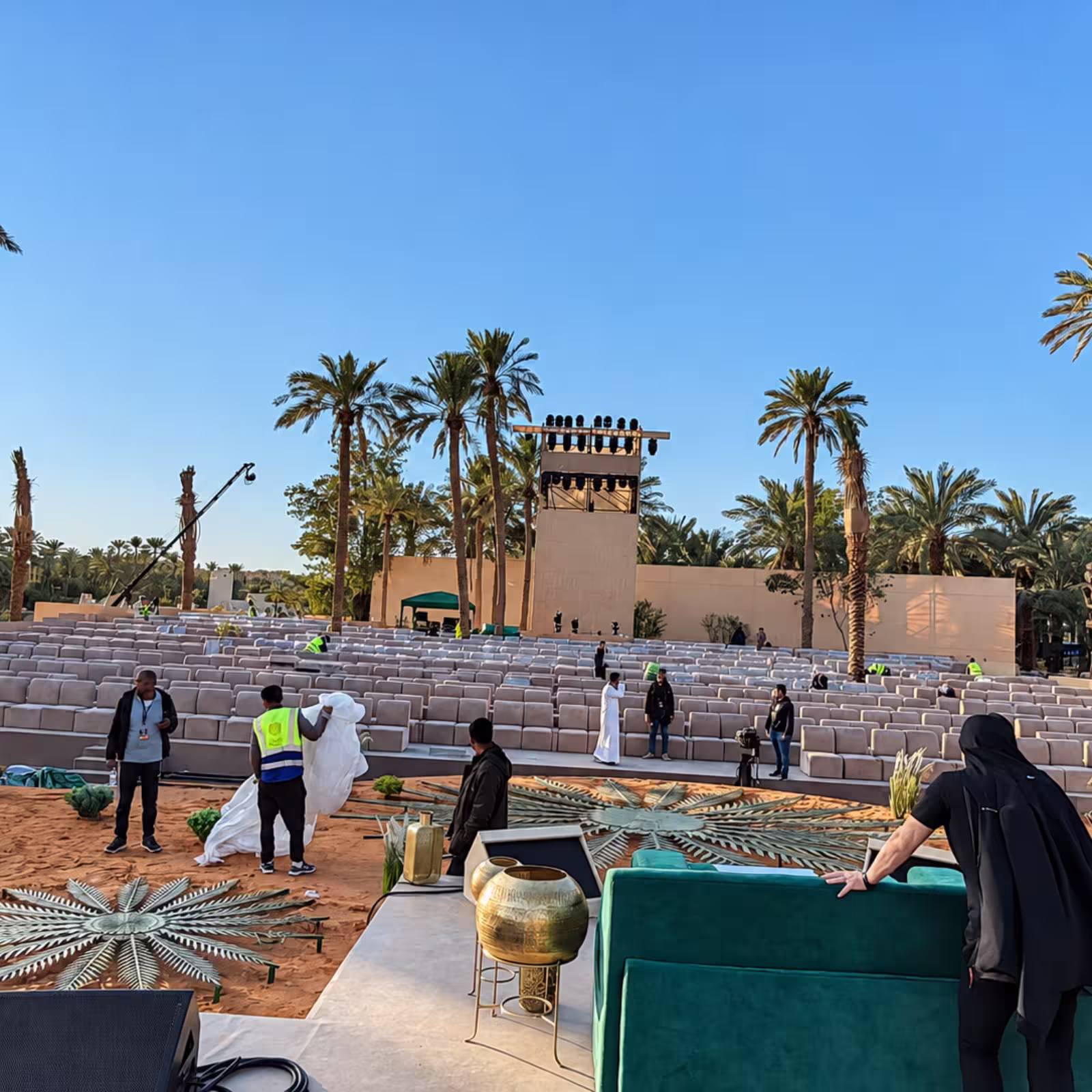 Outdoor event grandstand with rows of upholstered seating, palm trees, event lighting towers, and a decorated VIP or stage area in a landscaped venue.