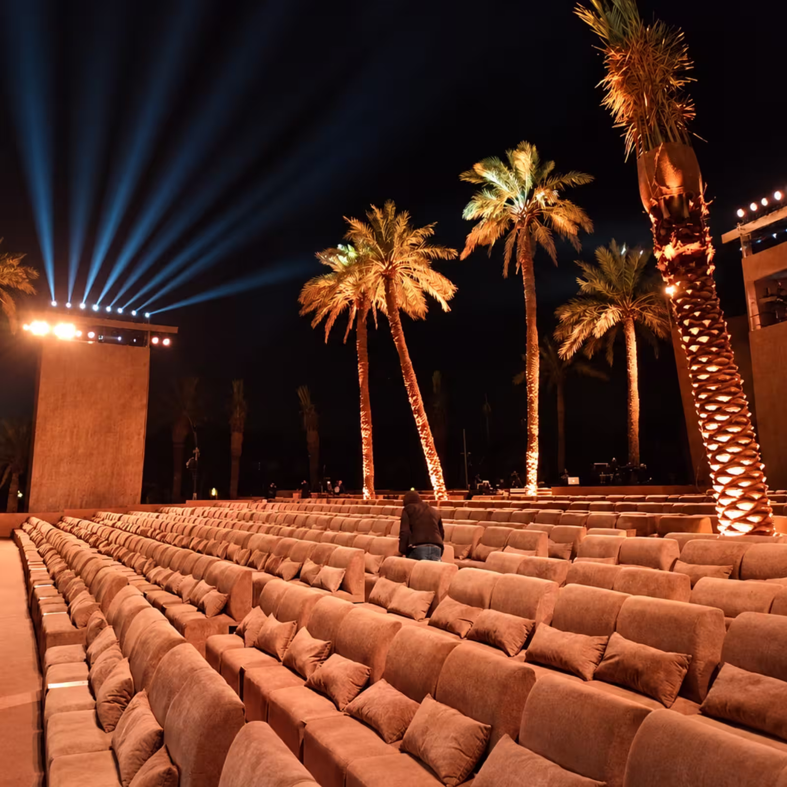Outdoor luxury event seating with rows of plush upholstered chairs, decorative cushions, illuminated palm trees, and dramatic stage lighting at a nighttime event venue.