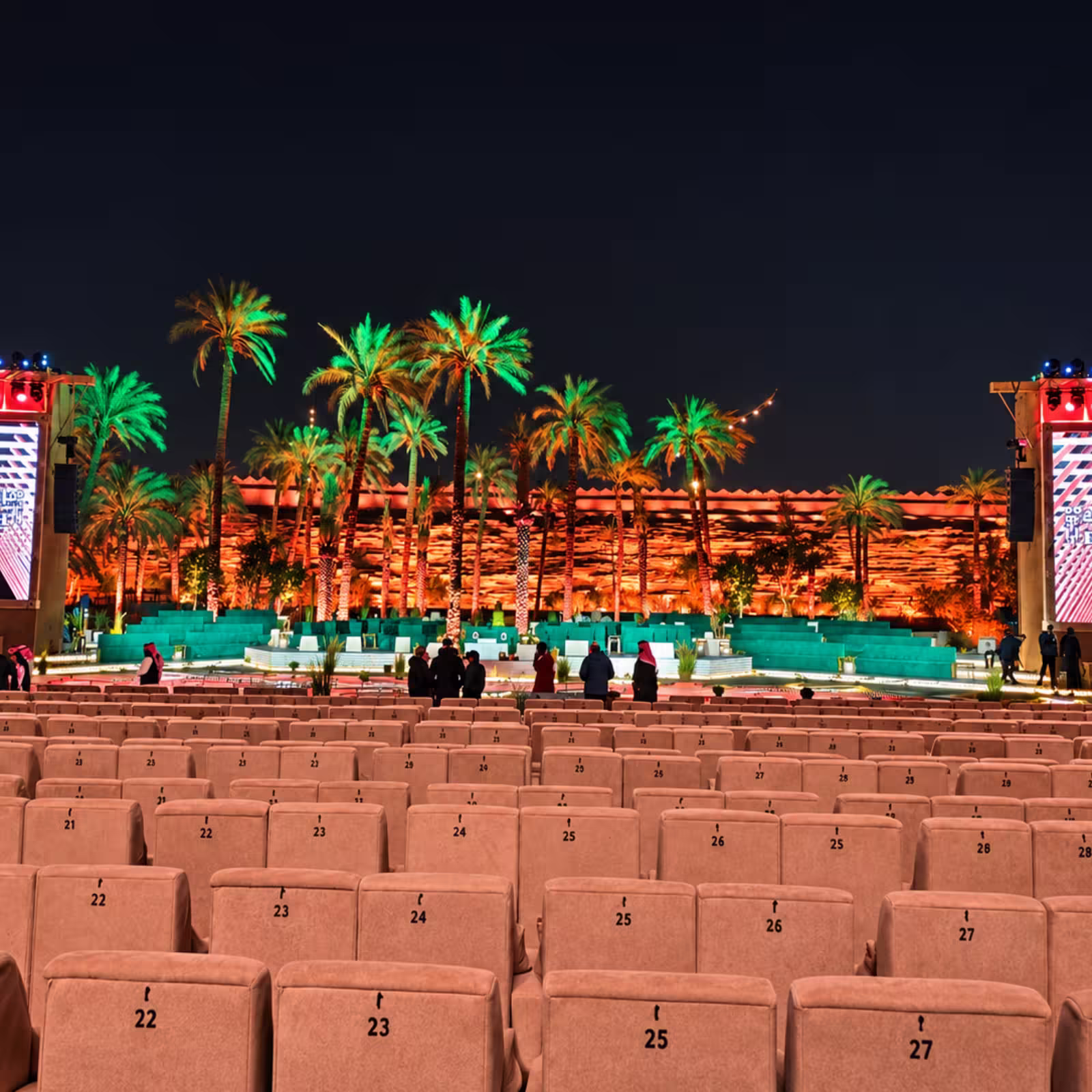 Outdoor luxury event seating with numbered upholstered chairs, illuminated palm trees, decorative stage seating, and vibrant nighttime event lighting.