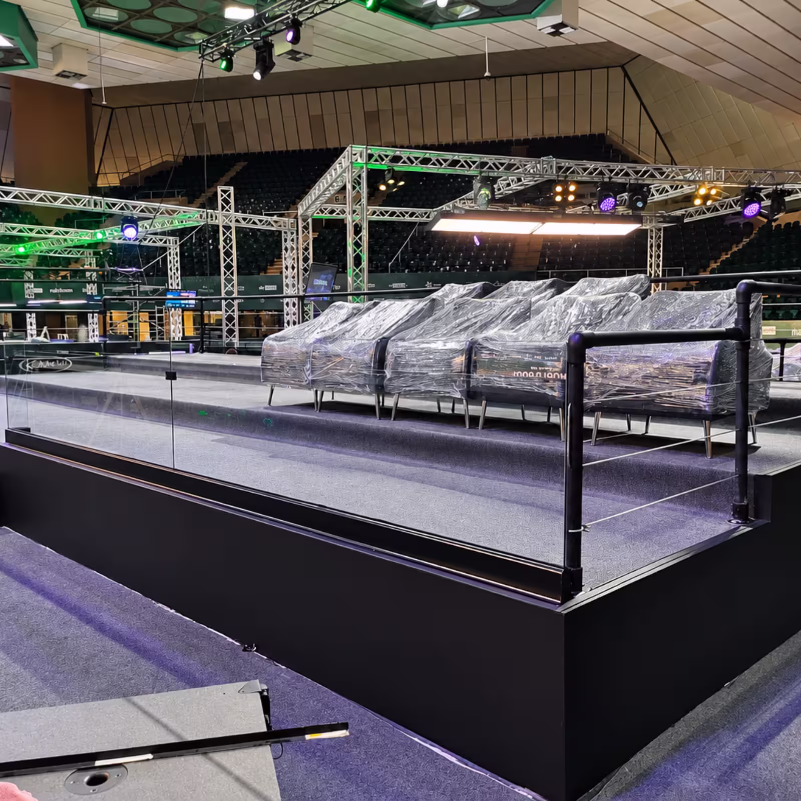 Indoor event seating area with wrapped lounge chairs, tiered platforms, stage lighting trusses, and a large arena-style venue prepared for an event.