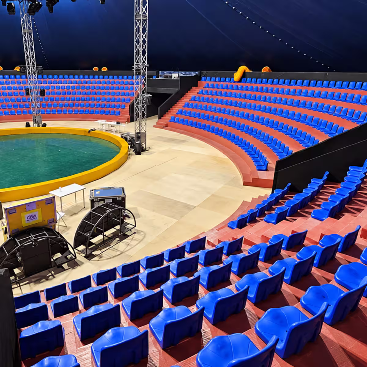 Temporary grandstand seating with rows of blue event chairs arranged around a circular arena, featuring tiered red seating platforms, event lighting trusses, and a central performance area.