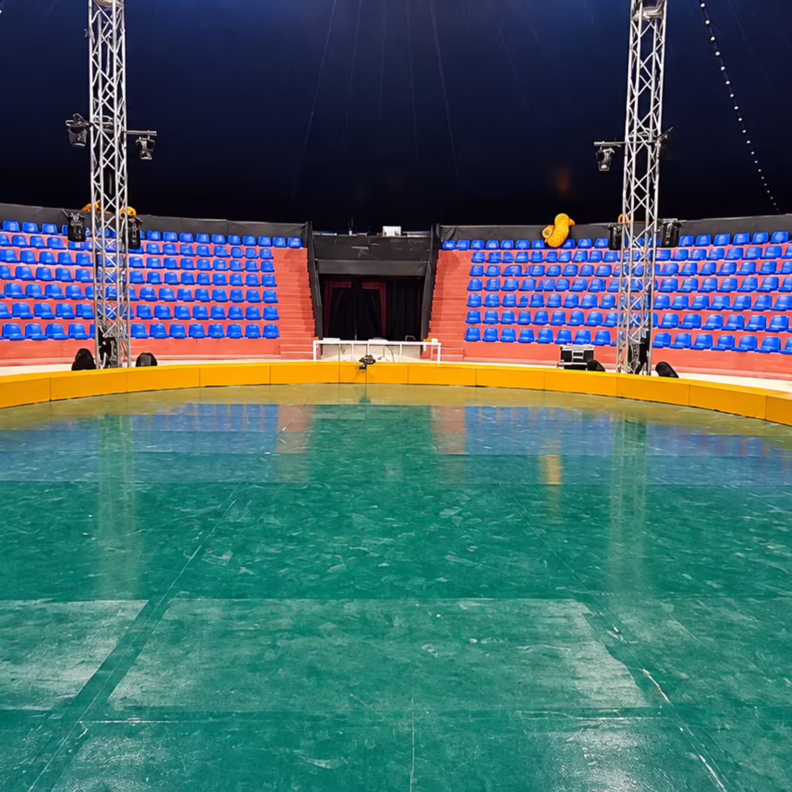 Grandstand seating surrounding a circular arena with blue spectator chairs, red tiered platforms, lighting trusses, and a central green performance floor.