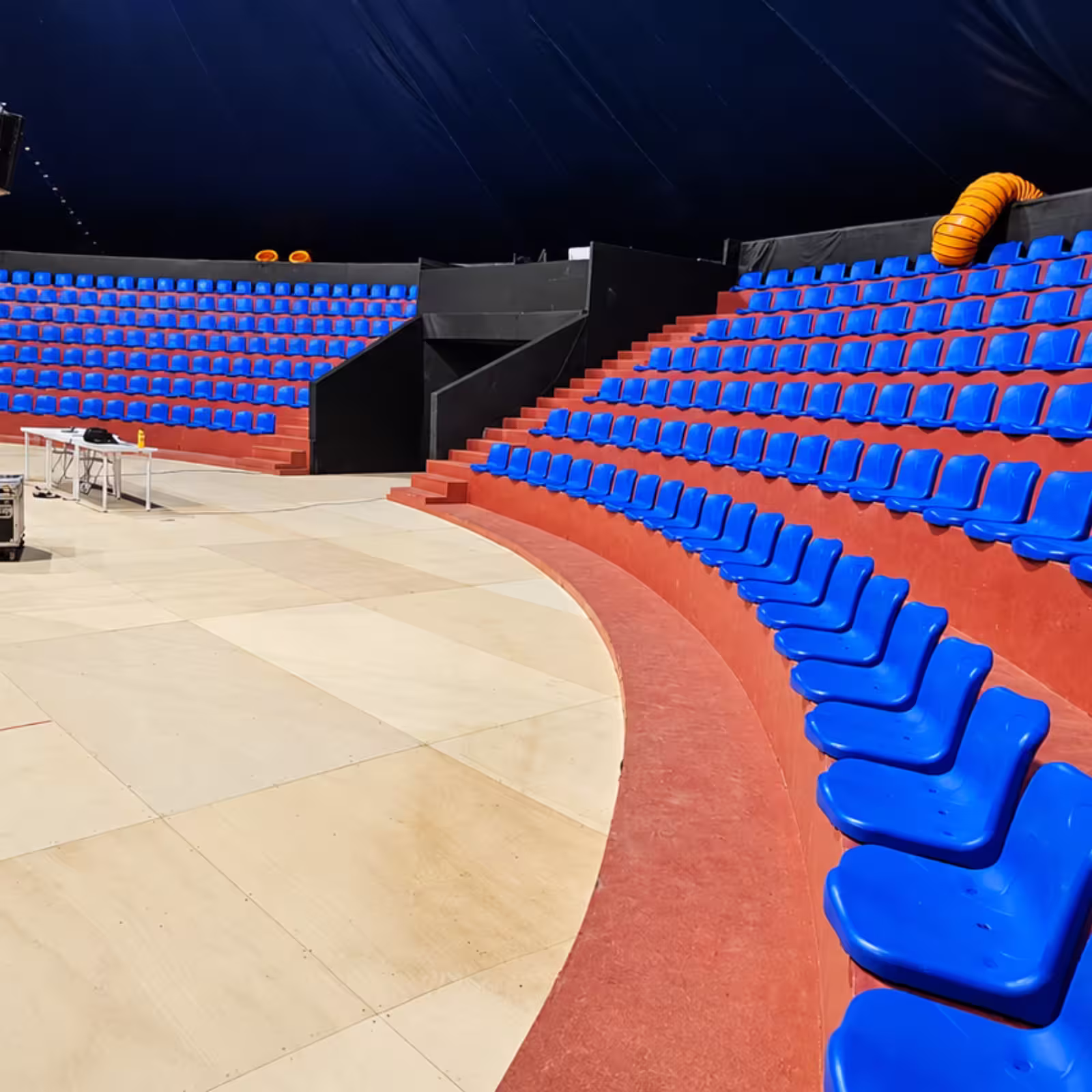 Temporary grandstand seating with rows of blue plastic chairs arranged in a curved arena layout on red tiered platforms.