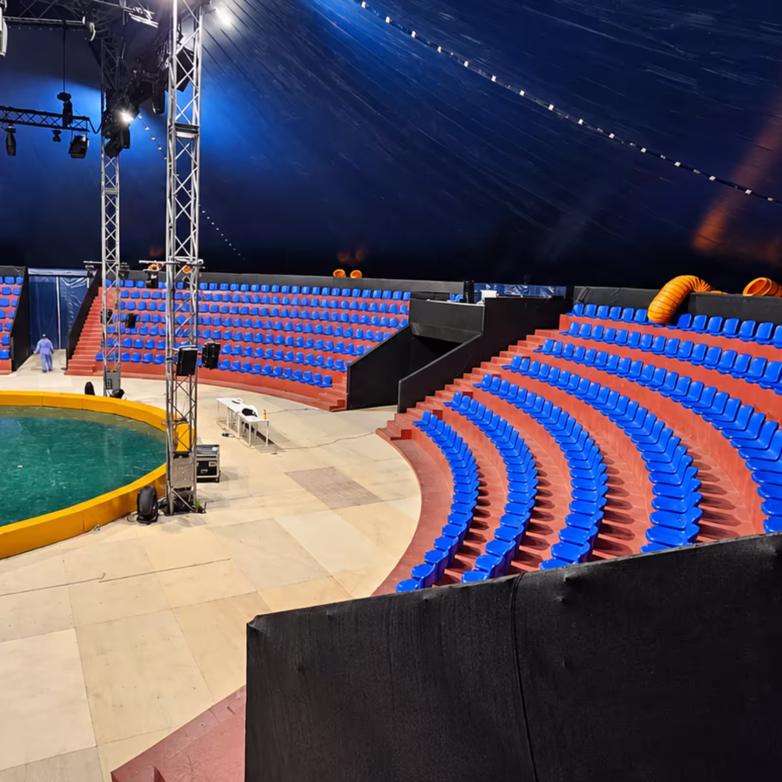 Temporary event grandstand with curved rows of blue seating, red tiered platforms, lighting trusses, and a circular performance arena beneath a large dark canopy.
