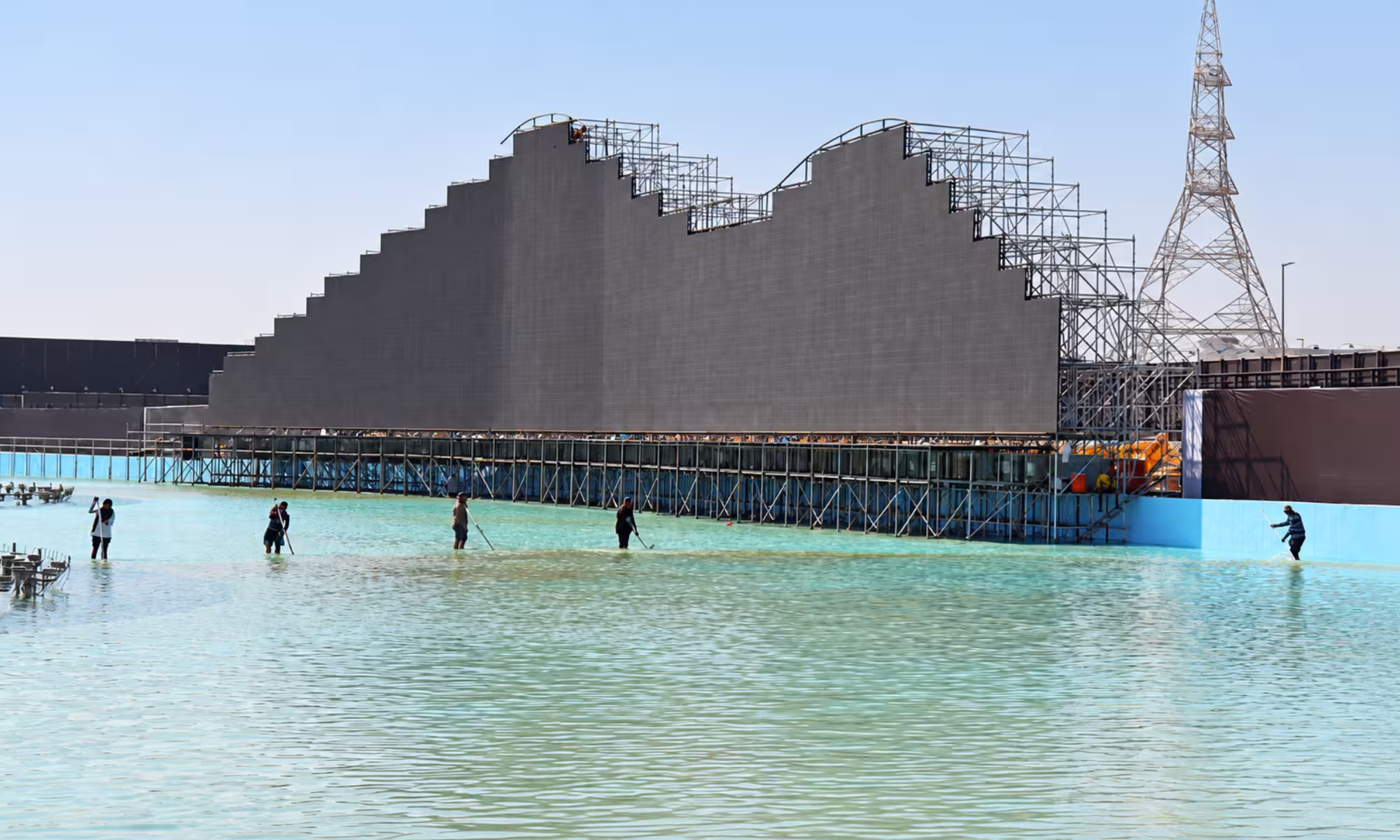 Temporary grandstand structure with large tiered seating platforms, steel support scaffolding, and a raised event viewing area built beside a large outdoor water or performance venue.
