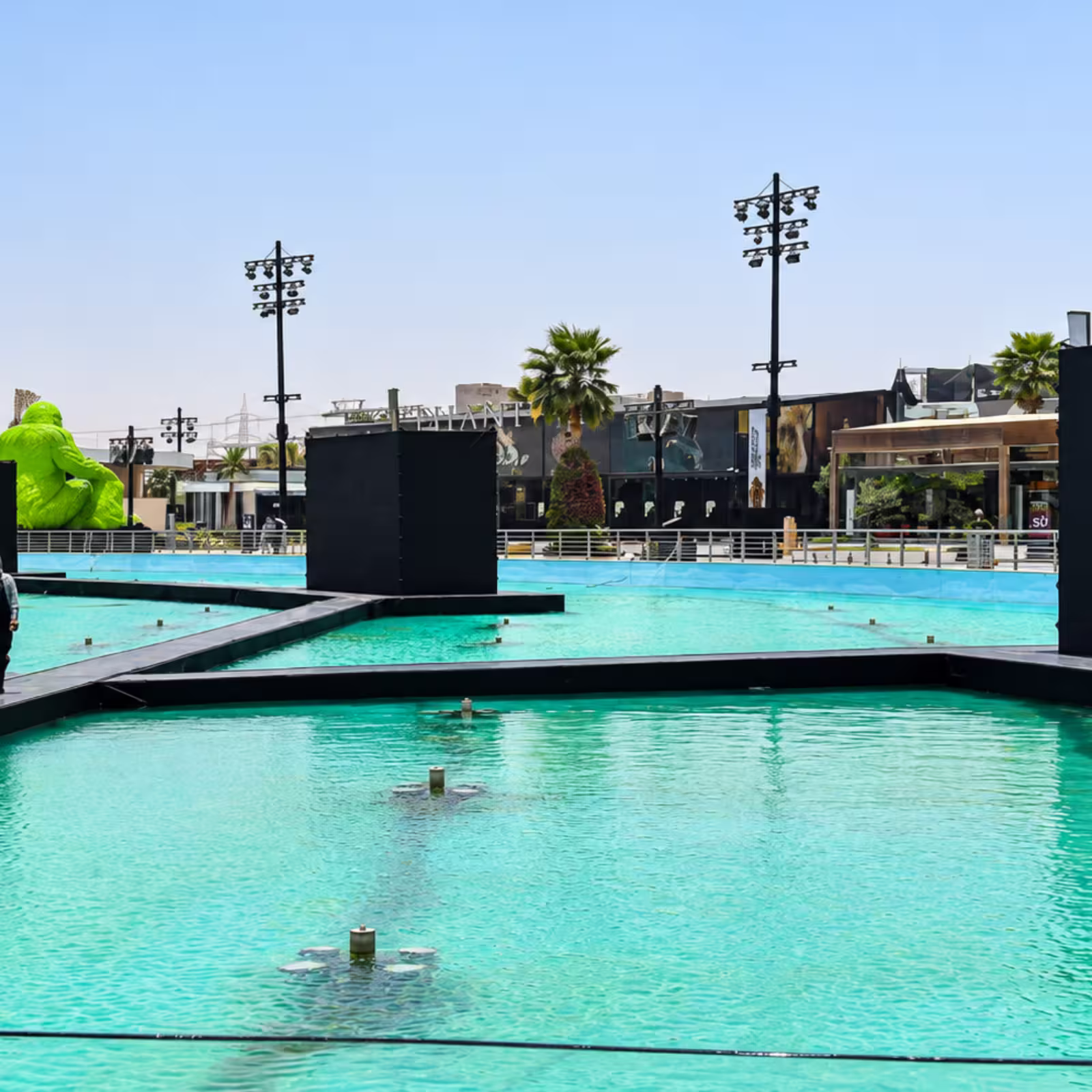 Outdoor event venue with a large turquoise pool, temporary black platforms and walkways, stage structures, event lighting towers, and palm trees surrounding the setup.
