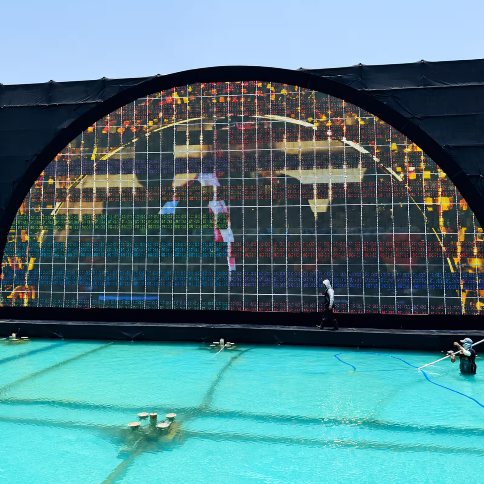 Large outdoor LED screen installation with an arched black frame positioned beside a turquoise pool, displaying a colorful digital visual during event setup.