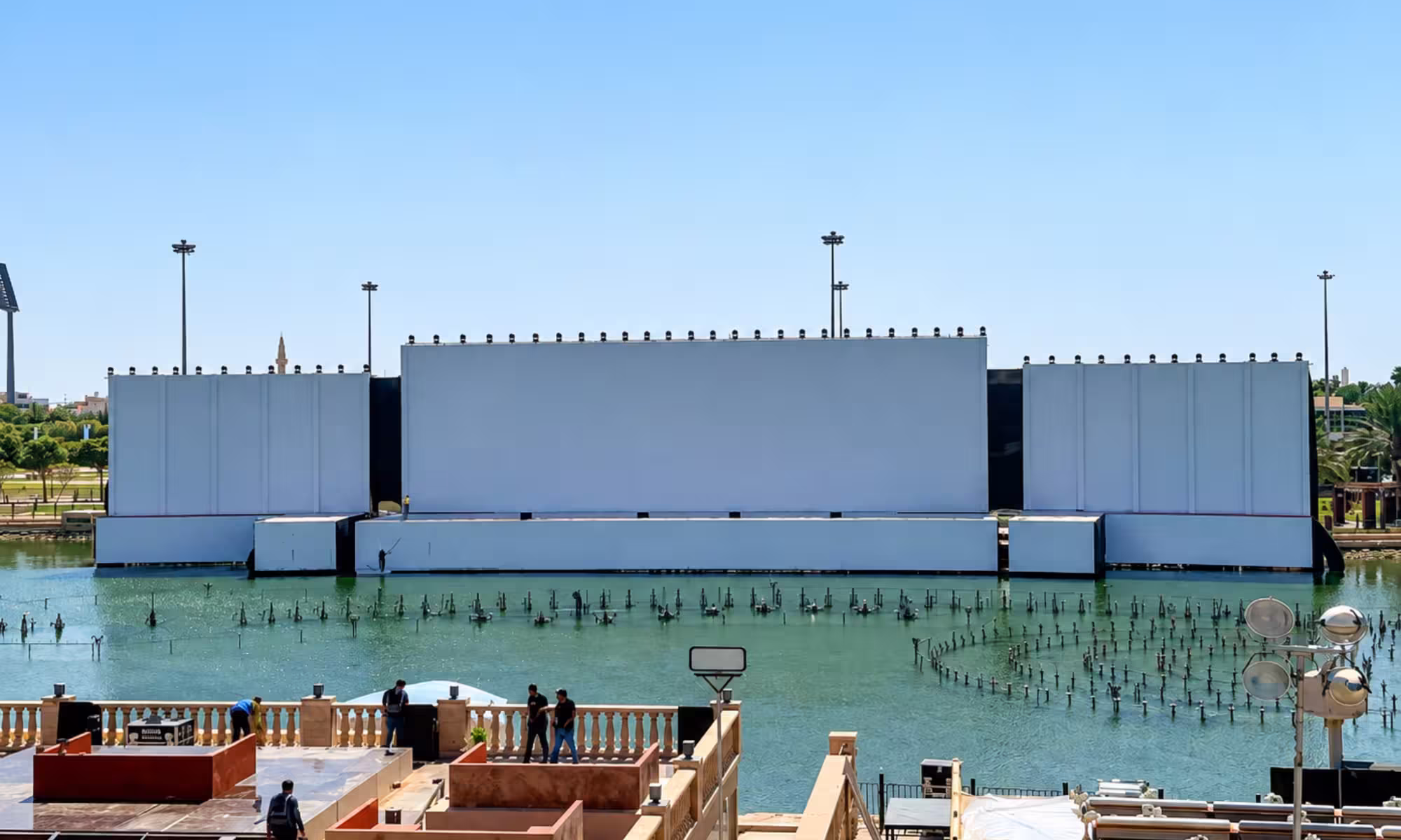 Large outdoor event-stage installation over a water feature, with a wide central screen structure, side panels, lighting fixtures, and event equipment surrounding the setup.