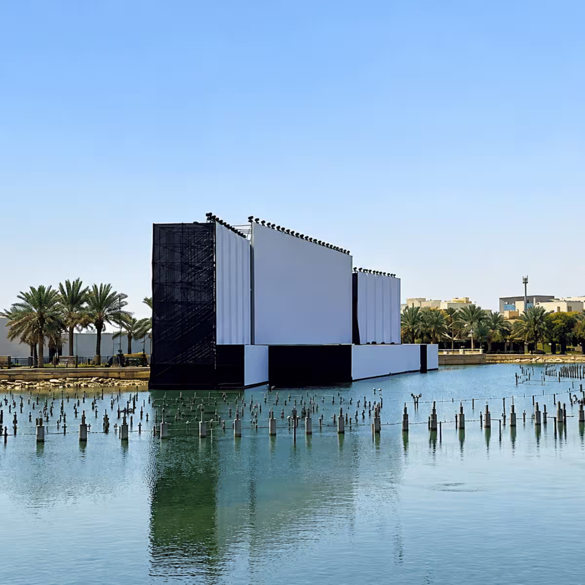 Large outdoor event-stage structure installed over a water feature, featuring a wide central screen, side screen panels, extensive stage lighting, and a large supporting truss/scaffolding structure.