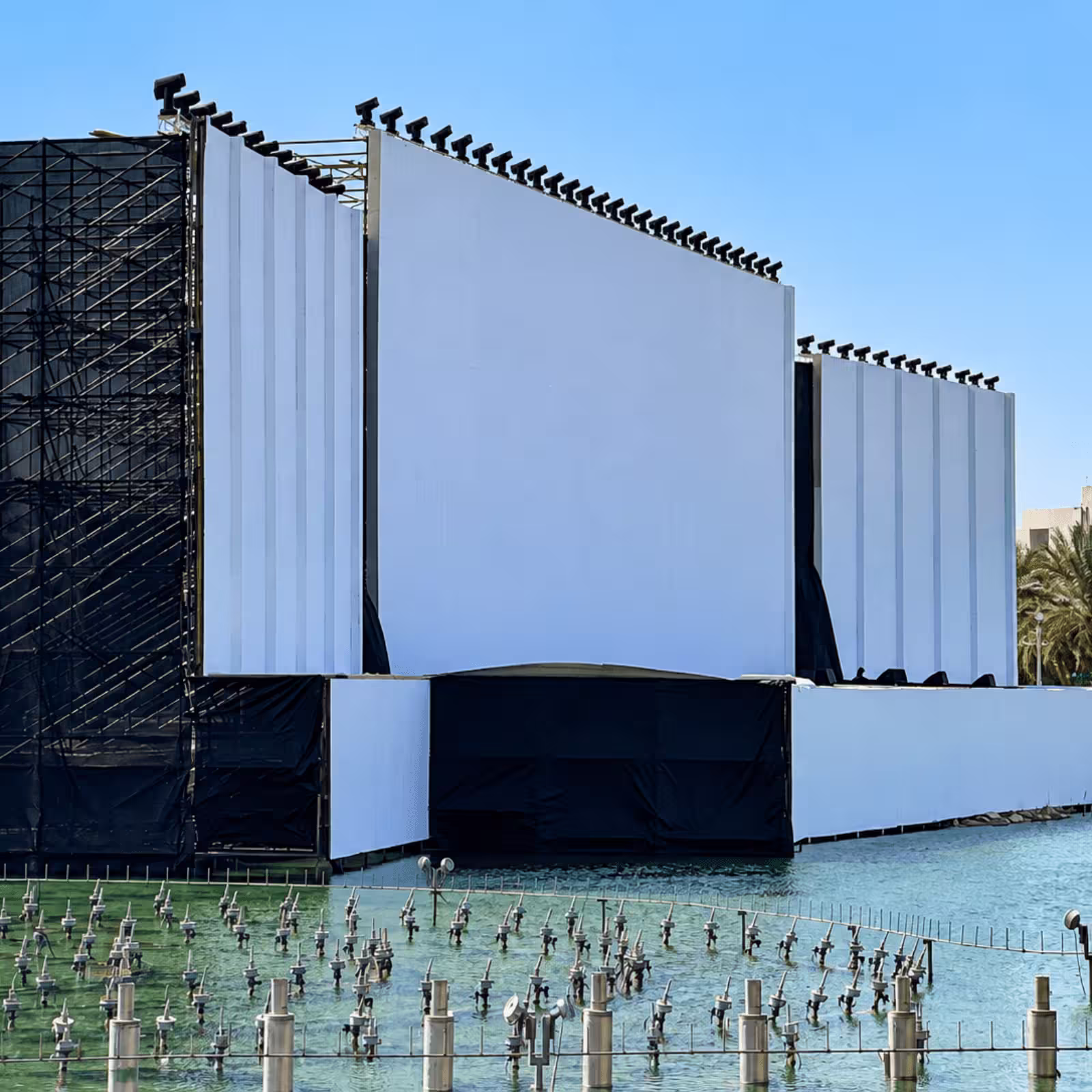 Large-scale outdoor event stage with a central projection/LED screen, matching side panels, perimeter stage lighting, heavy scaffolding, and water-based event infrastructure.