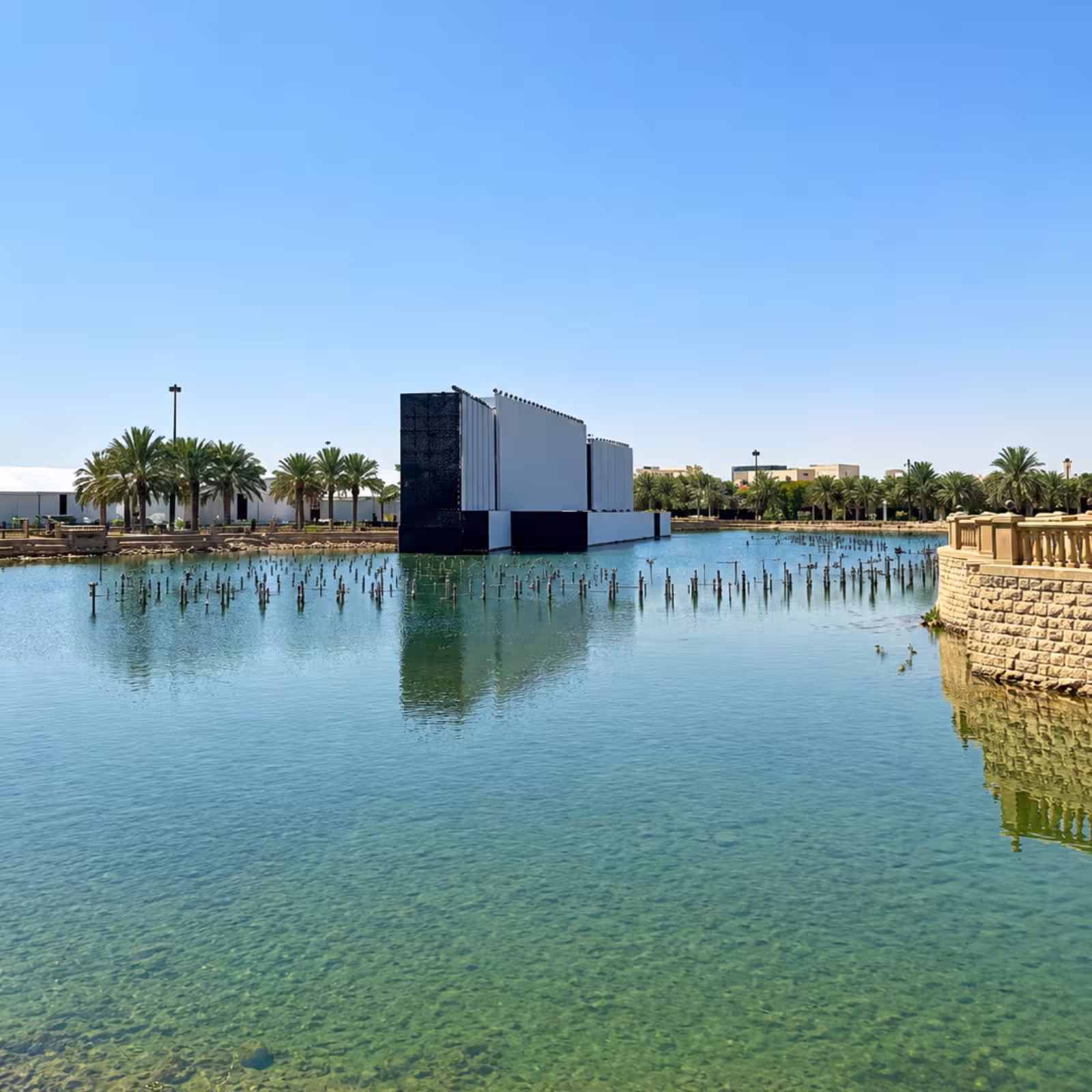 Wide view of a large outdoor event-stage installation positioned on a water feature, surrounded by palm trees and landscaped grounds, with a prominent central stage structure and extensive event infrastructure.