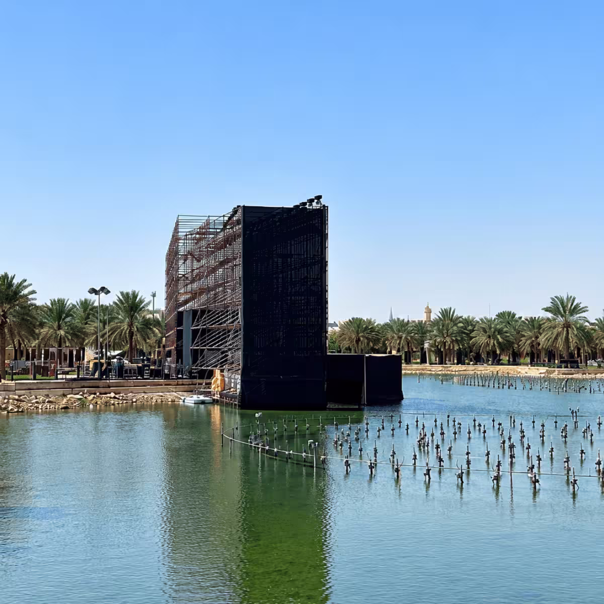 Large-scale event-stage installation positioned over a water feature, showing a substantial black scaffolding structure, stage framework, lighting equipment, and surrounding palm-lined grounds.
