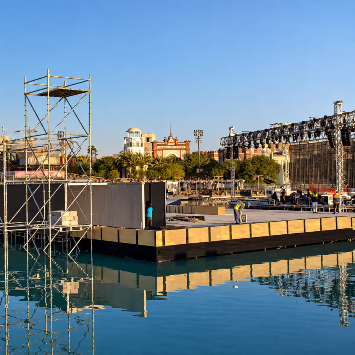Outdoor floating event stage setup with scaffolding, truss lighting, LED screens, and production equipment over a waterfront venue.