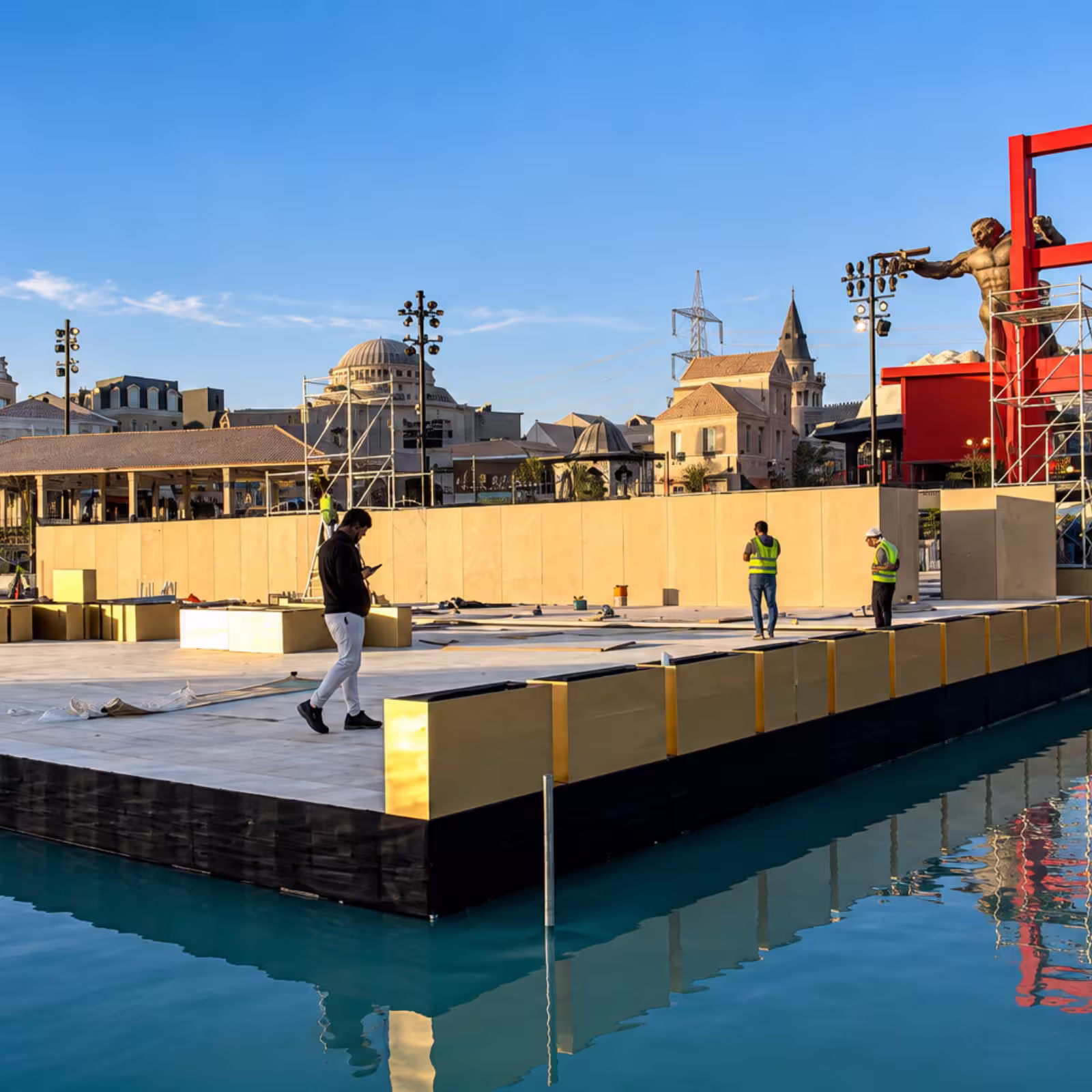 Floating event stage under construction over a waterfront, with lighting towers, scaffolding, stage panels, and event crew.