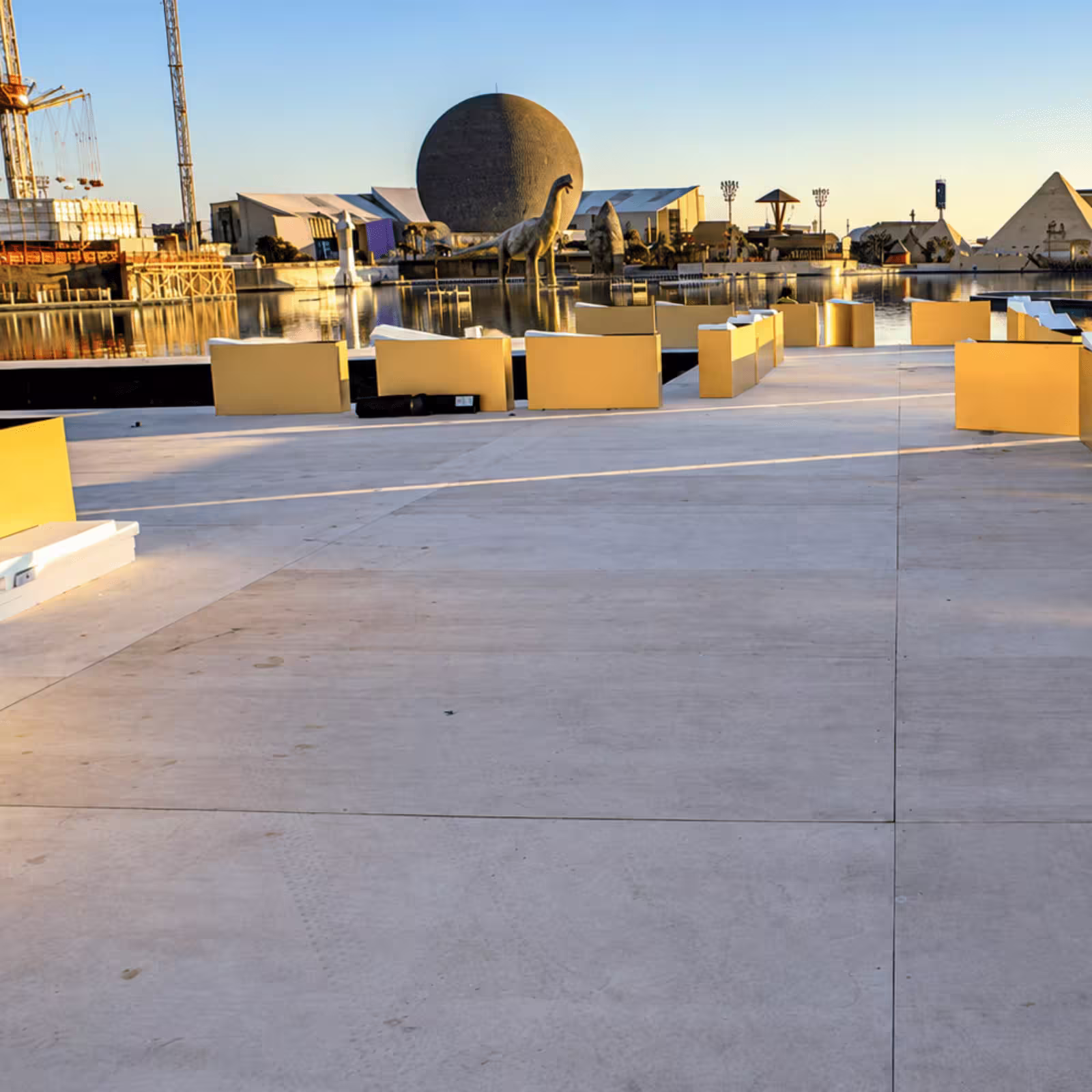 Outdoor event platform with gold seating barriers beside a waterfront venue, featuring large sculptures, temporary structures, and event infrastructure.