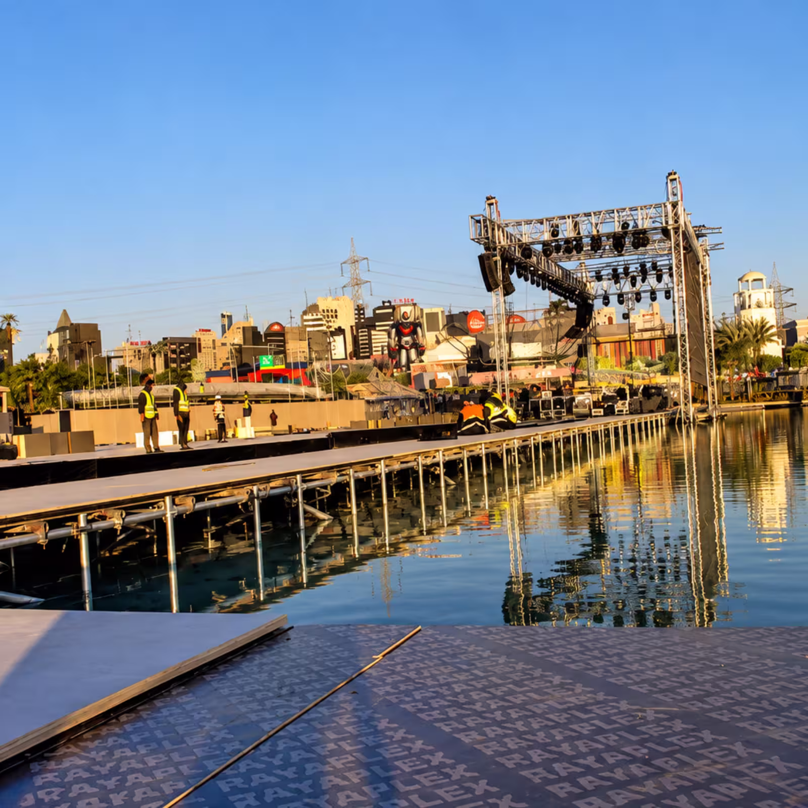 Temporary event stage and waterfront platform under construction, with lighting truss, workers, and city buildings in the background.