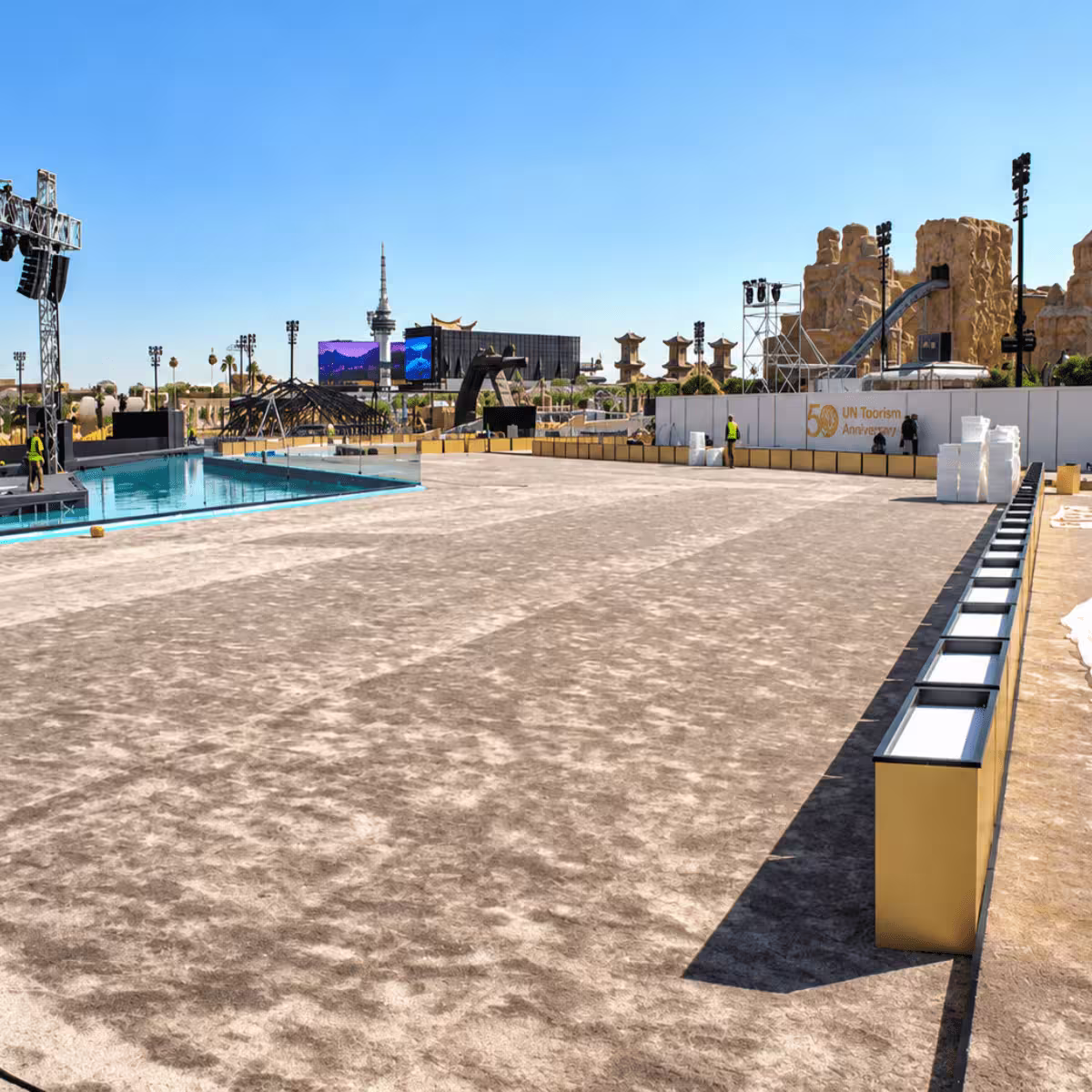 Large outdoor event venue under construction with temporary stages, lighting towers, screens, barriers, and a pool area.