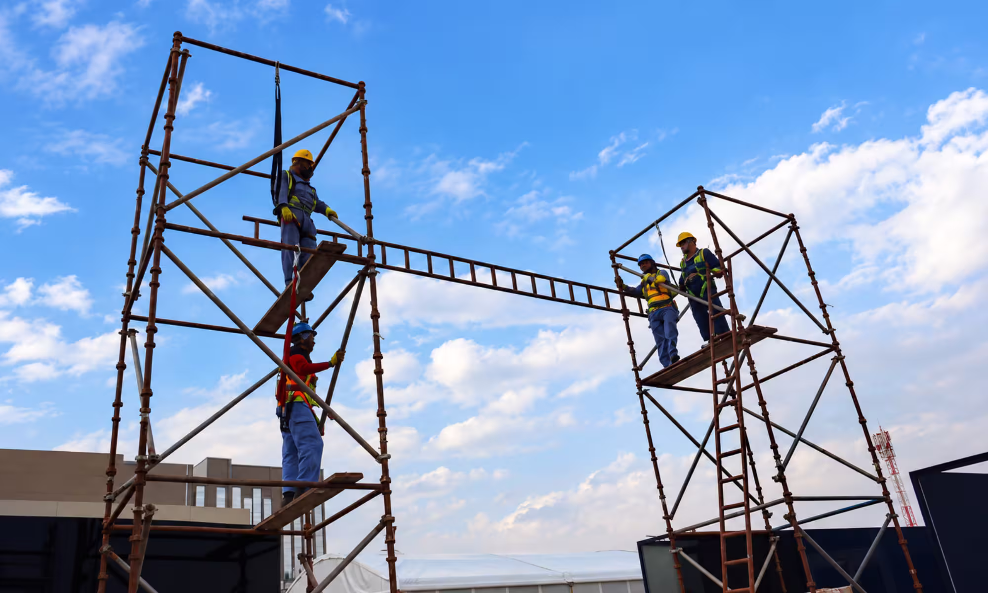 Workers in safety gear assembling a tall metal scaffolding structure at an outdoor event site.