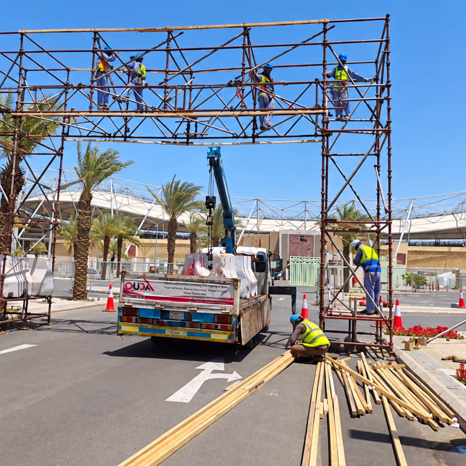 Workers assembling a large metal scaffolding structure at an outdoor event venue, with a truck, crane, and construction materials on site.