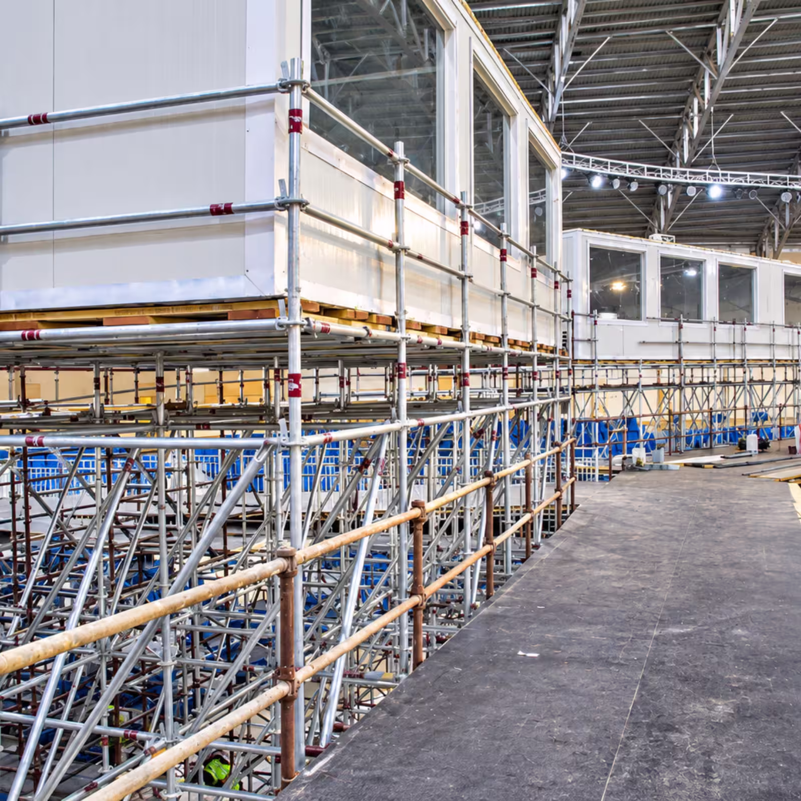 Indoor event structure with metal scaffolding supporting elevated platforms and temporary rooms inside a large exhibition venue.