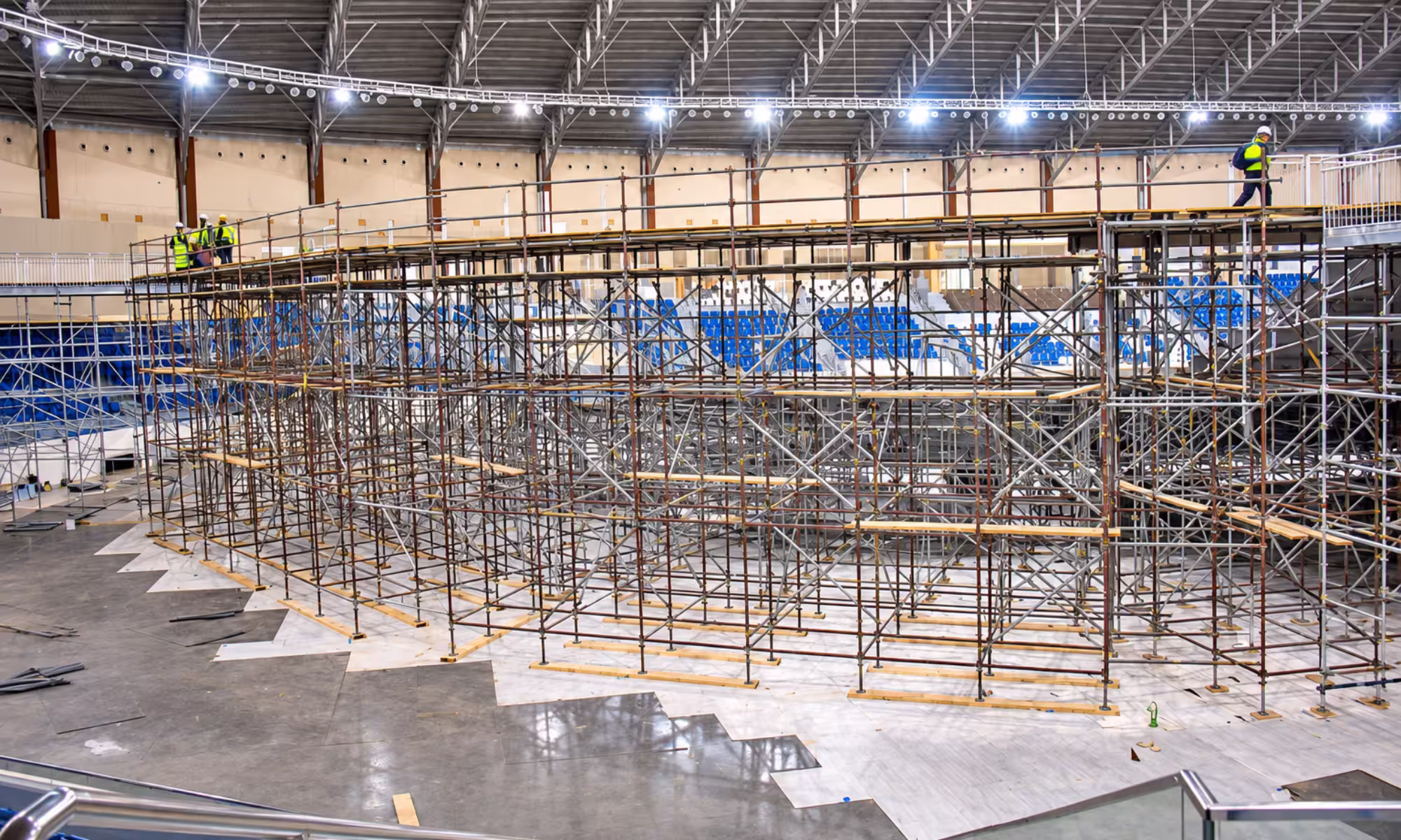 Large indoor event venue with extensive multi-level scaffolding and workers installing elevated platforms and seating areas.