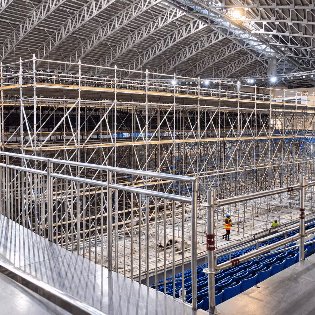 Indoor event venue with extensive multi-level scaffolding, elevated platforms, blue seating, and workers during construction setup.