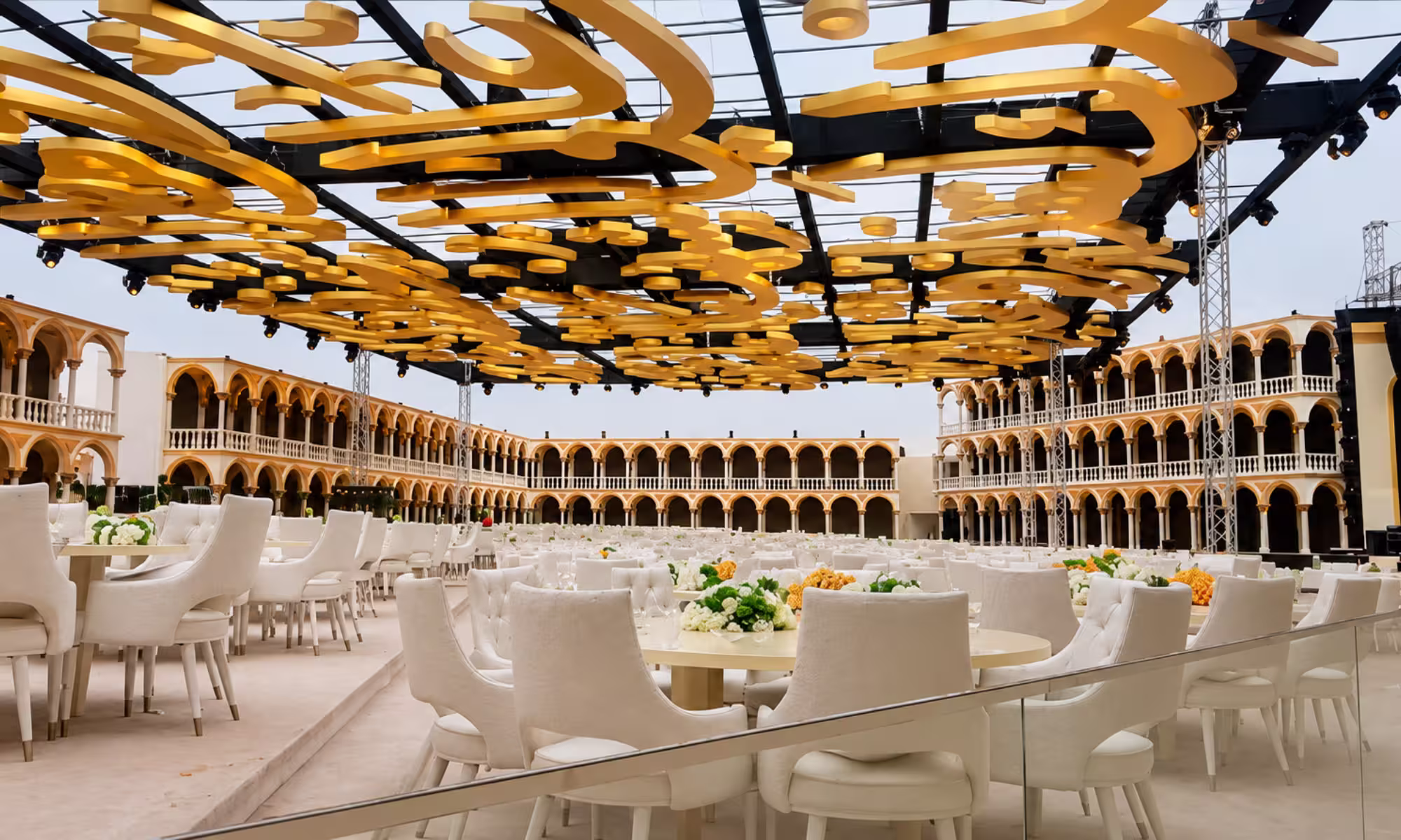 Elegant outdoor event venue with white banquet seating, floral table settings, and an ornate golden overhead decorative installation.