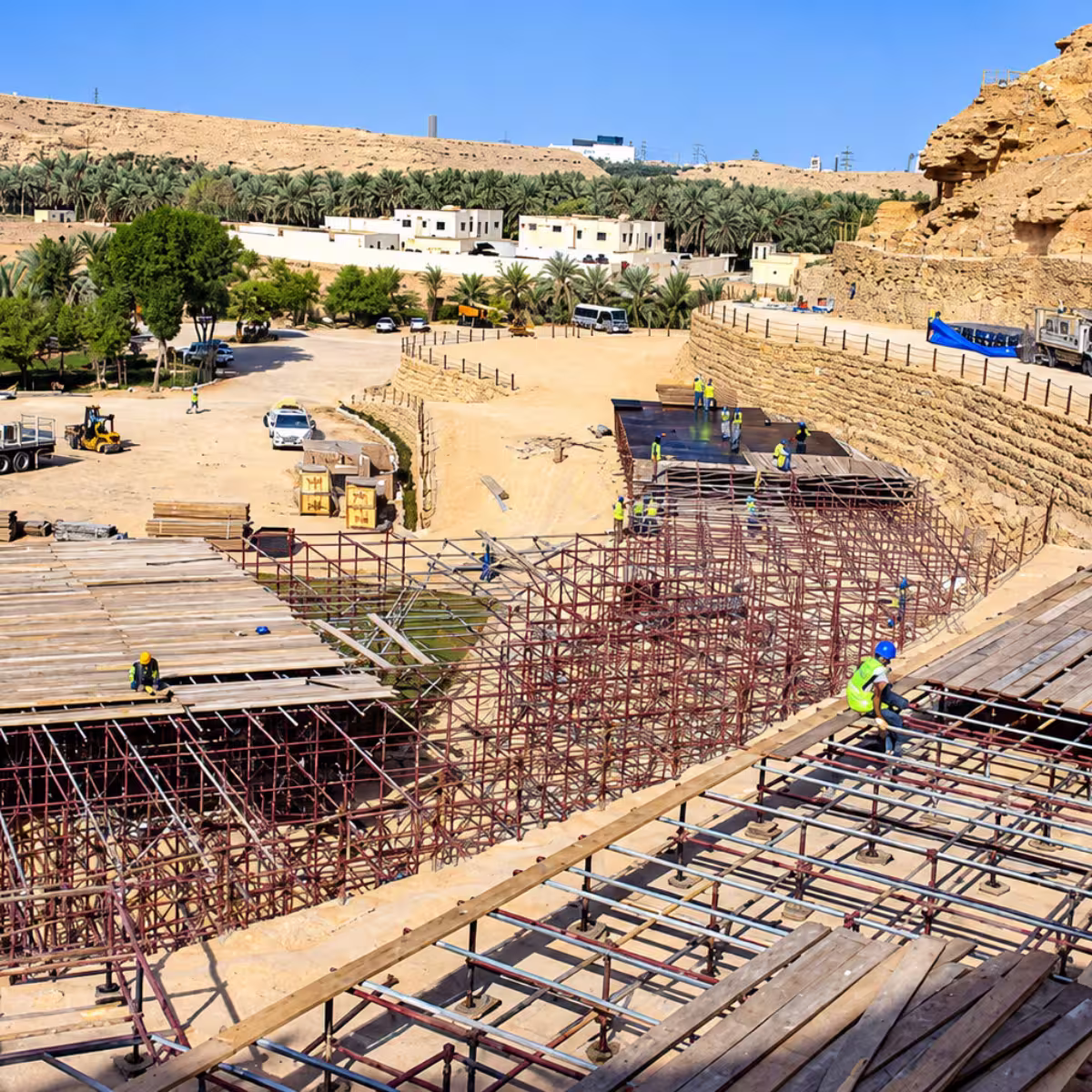 Large-scale outdoor construction project with workers assembling extensive scaffolding and elevated platforms in a desert oasis landscape.
