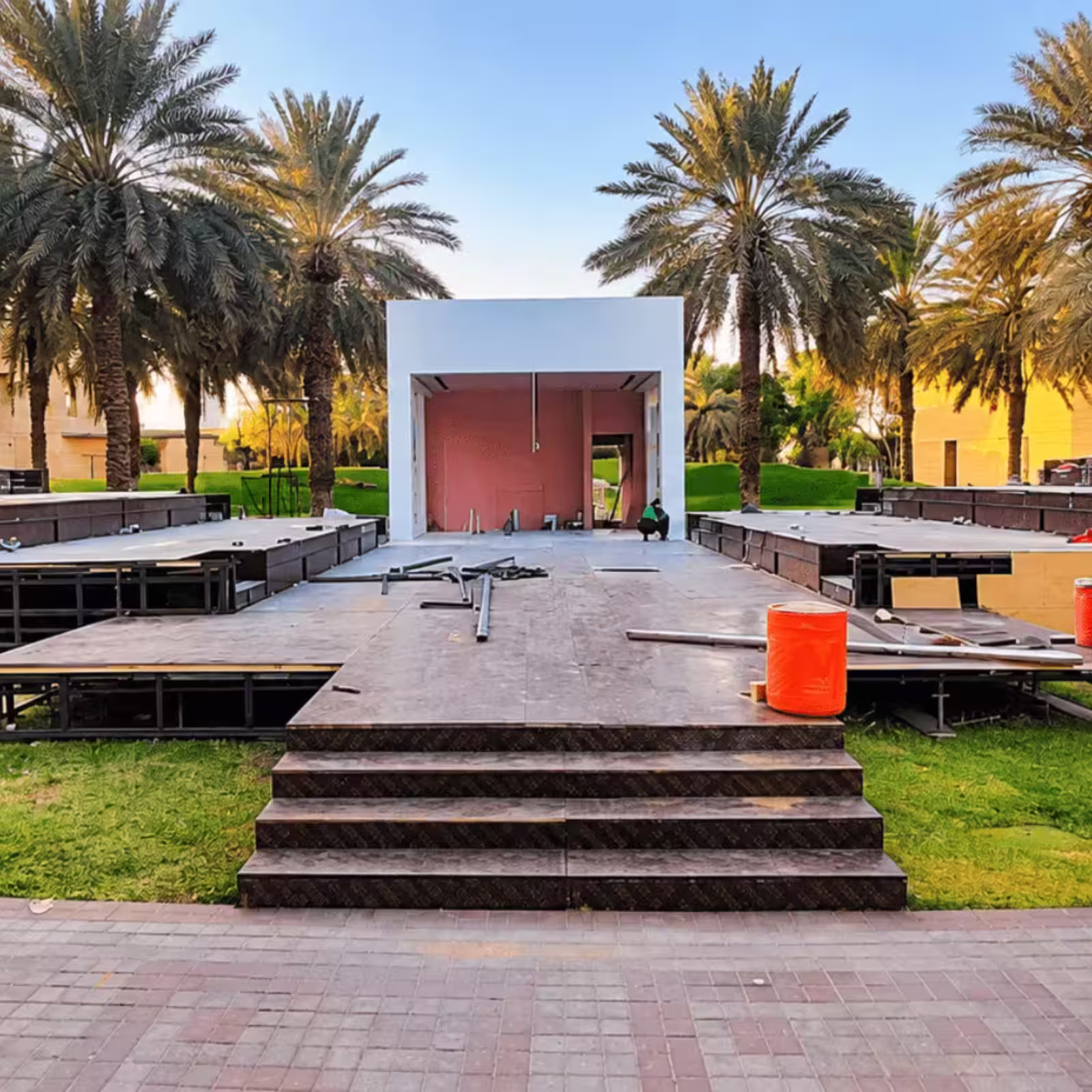 Outdoor event stage platform under construction in a landscaped venue with palm trees and a central stage entrance.