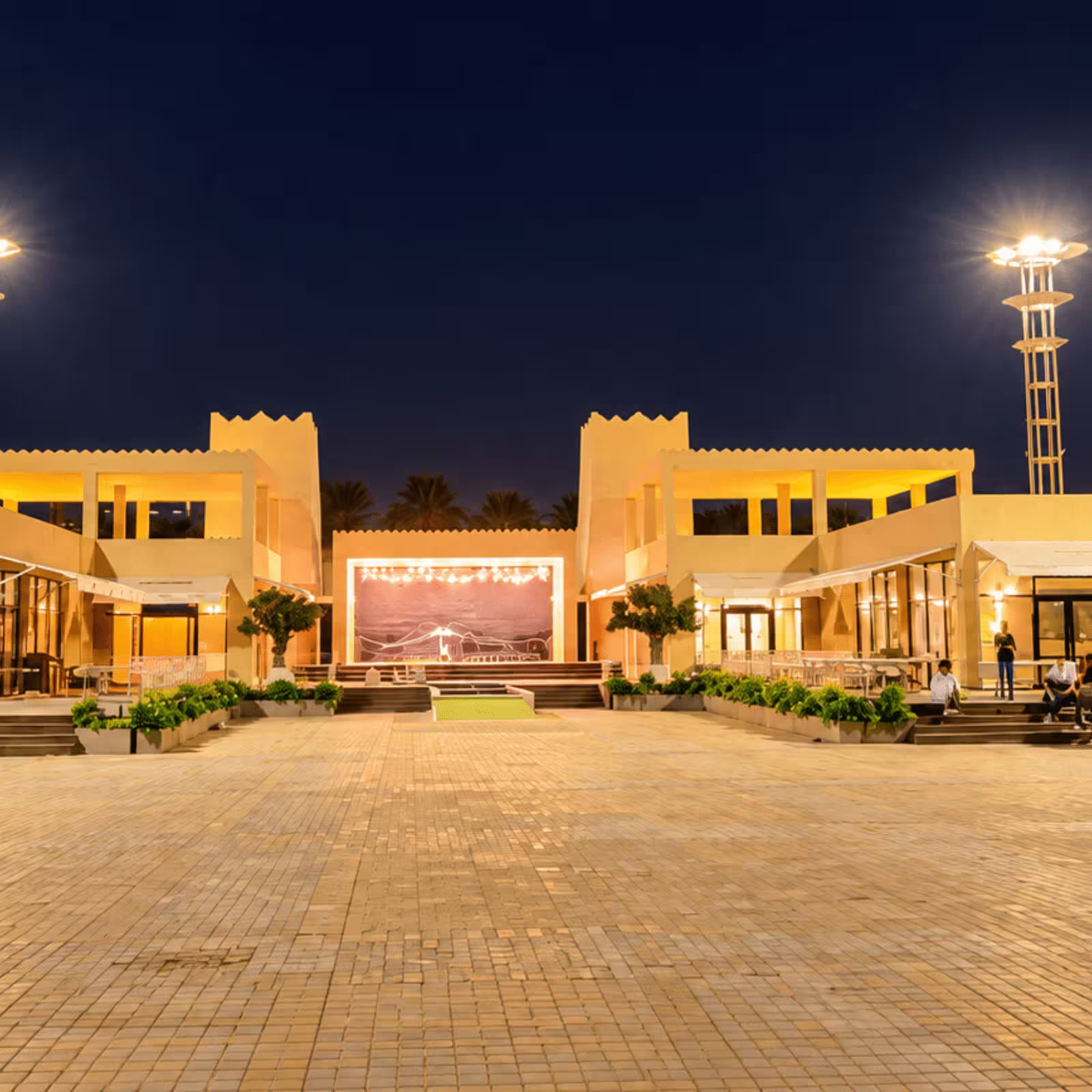 Outdoor event venue at night with a central stage, illuminated traditional-style buildings, decorative landscaping, and tall event lighting towers.
