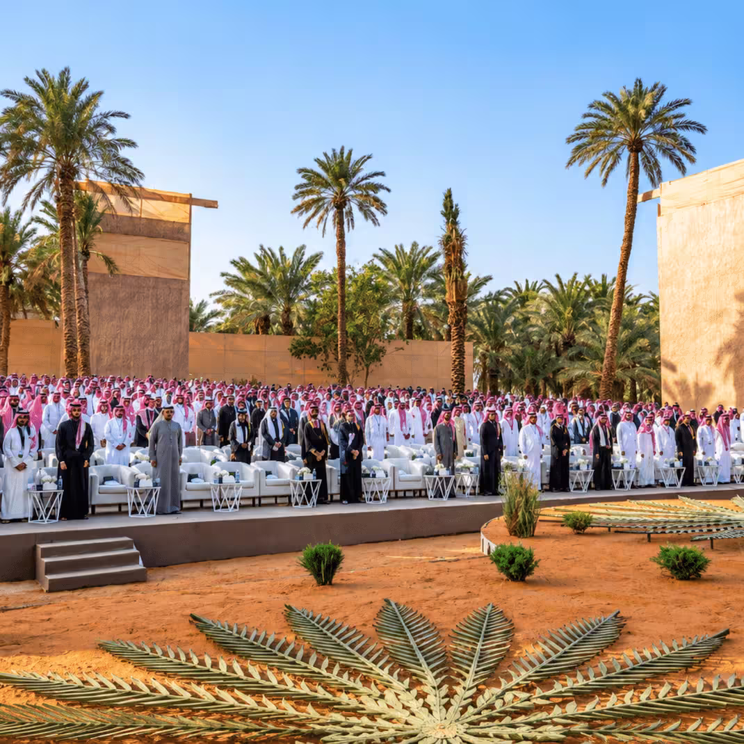 Outdoor Saudi Arabian event with a large seated audience, white lounge seating, palm trees, and desert-inspired landscaping.