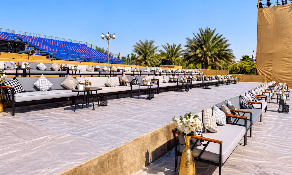 Outdoor event seating area with white lounge sofas, patterned cushions, floral arrangements, palm trees, and blue spectator stands.