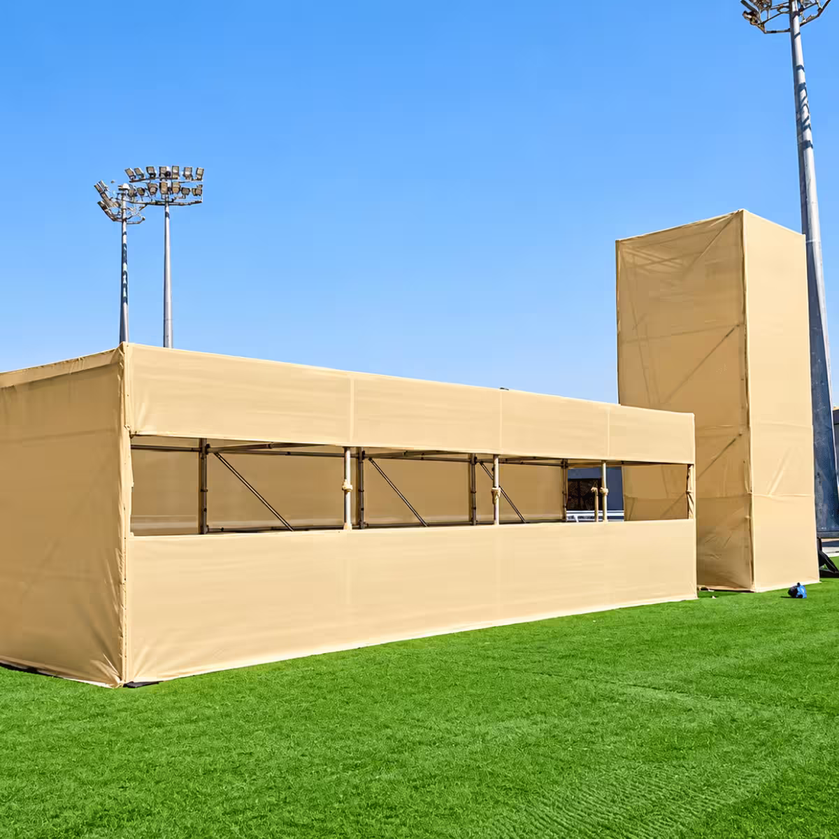 Outdoor event structure with beige fabric-covered scaffolding, integrated lighting towers, and a green grass field.