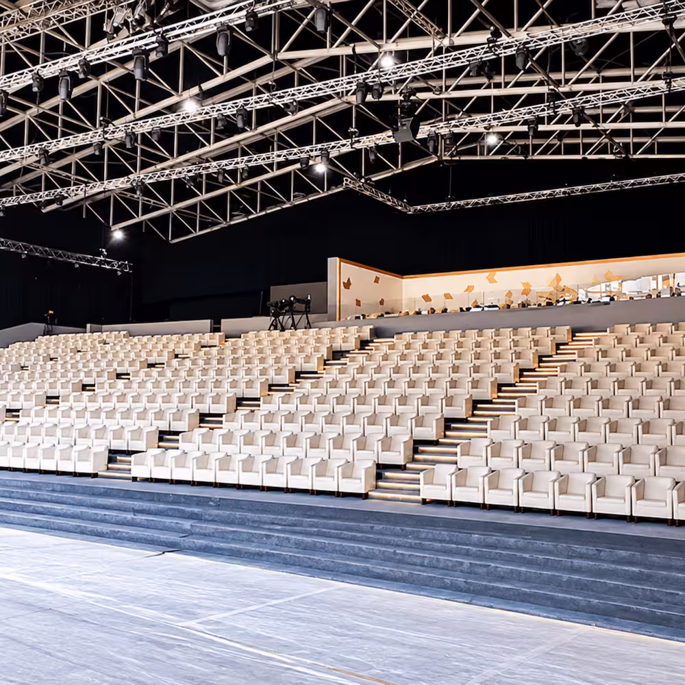 Indoor event venue with tiered white seating, illuminated stairways, and extensive overhead stage lighting trusses.